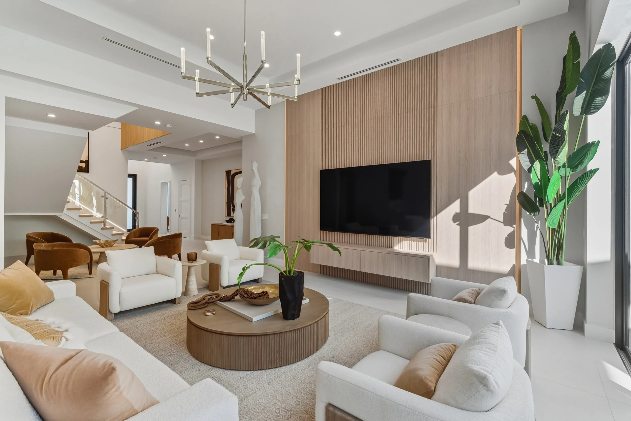 Modern living room with white and beige furniture, a large TV on a wood-paneled wall, tall green plants, and a contemporary chandelier.
