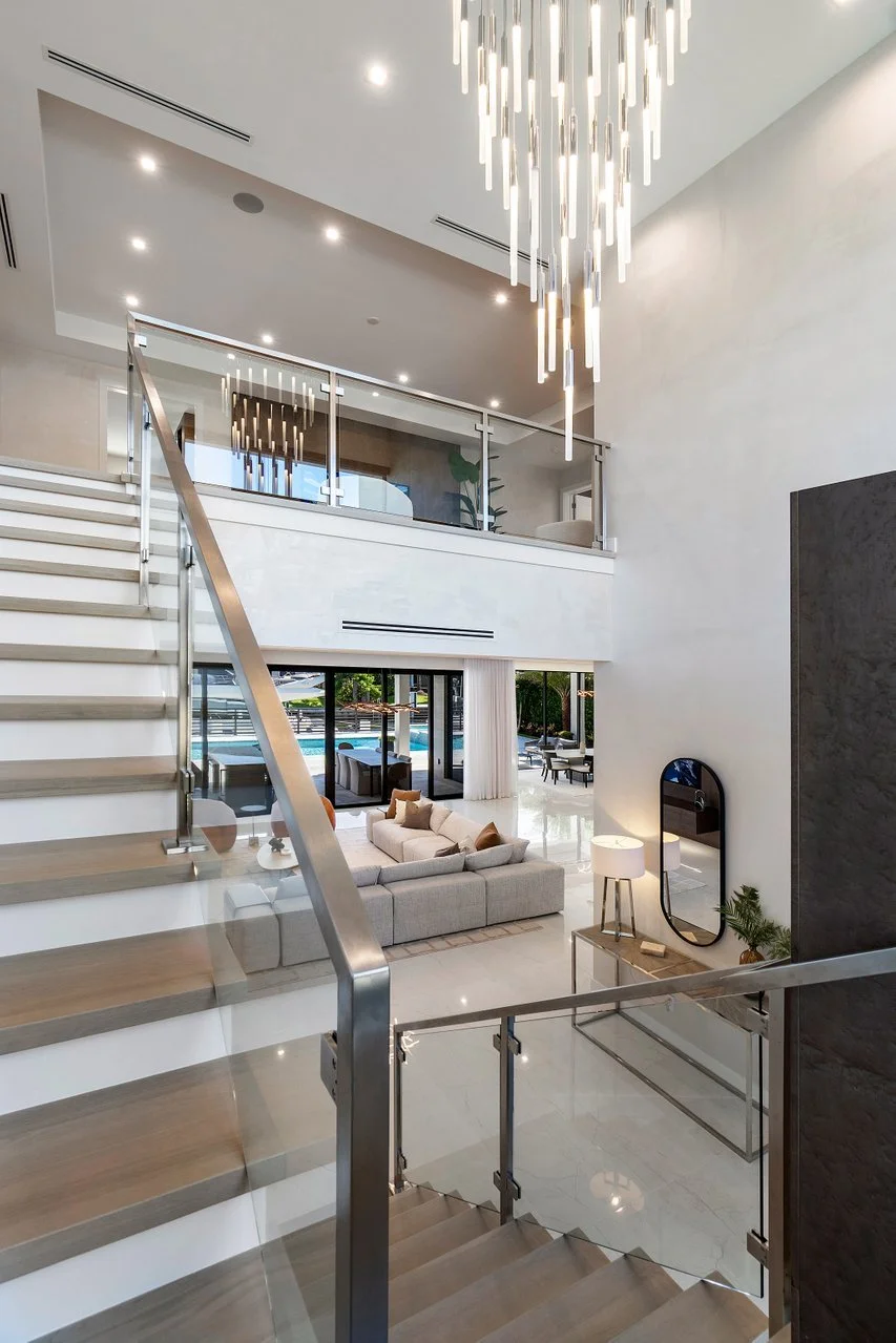 Interior view of a modern, spacious home with a staircase leading to an upper level, a sitting area with a sofa, a chandelier hanging from the ceiling, and large sliding glass doors opening to an outdoor pool area.