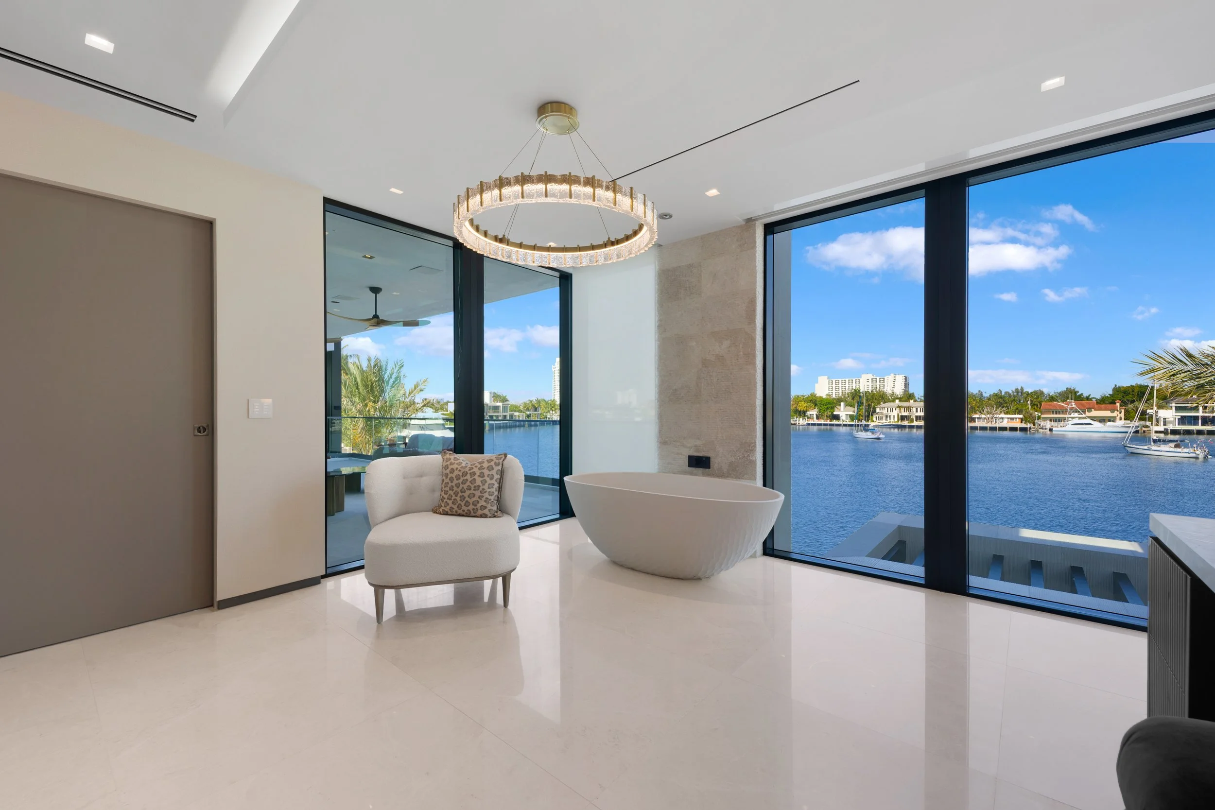 Modern living space with large floor-to-ceiling windows overlooking water and boats, featuring a white armchair with a patterned pillow, a curved bathtub, and a chandelier on a white ceiling.