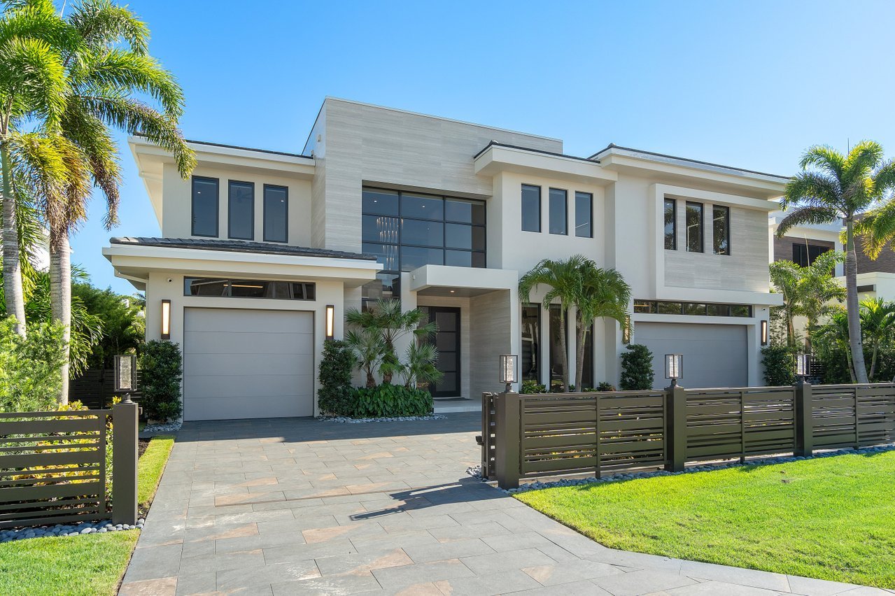 Modern two-story house with a white exterior, large windows, and two garages, surrounded by palm trees and a landscaped yard under a clear blue sky.
