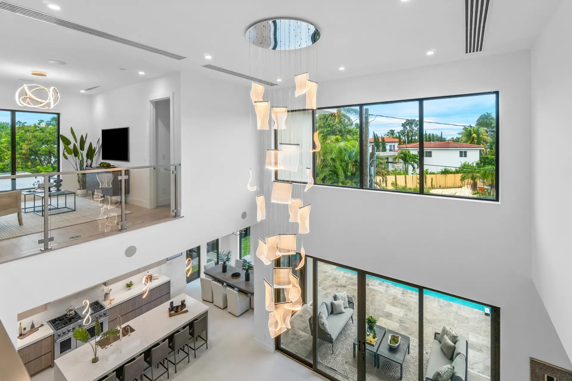 Interior view of a modern, two-story house living area with large windows showing trees and neighboring houses, featuring a pendant light fixture, white walls, and contemporary furniture.
