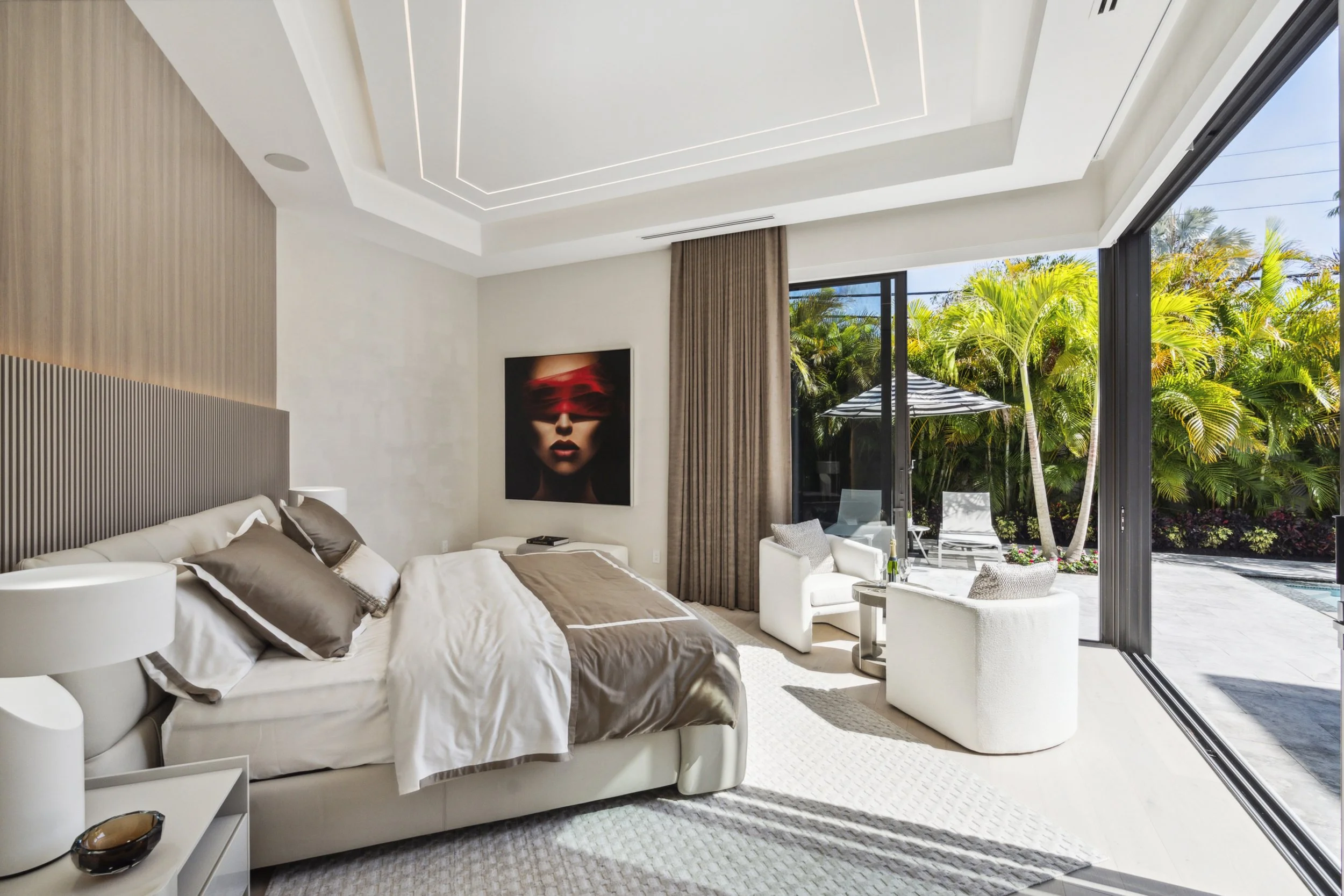 A modern bedroom with a large bed, beige and brown bedding, and white bedside tables. There is a painting of a woman with red glasses on the wall, and large windows with sliding glass doors opening to a patio with outdoor furniture and lush green trees.