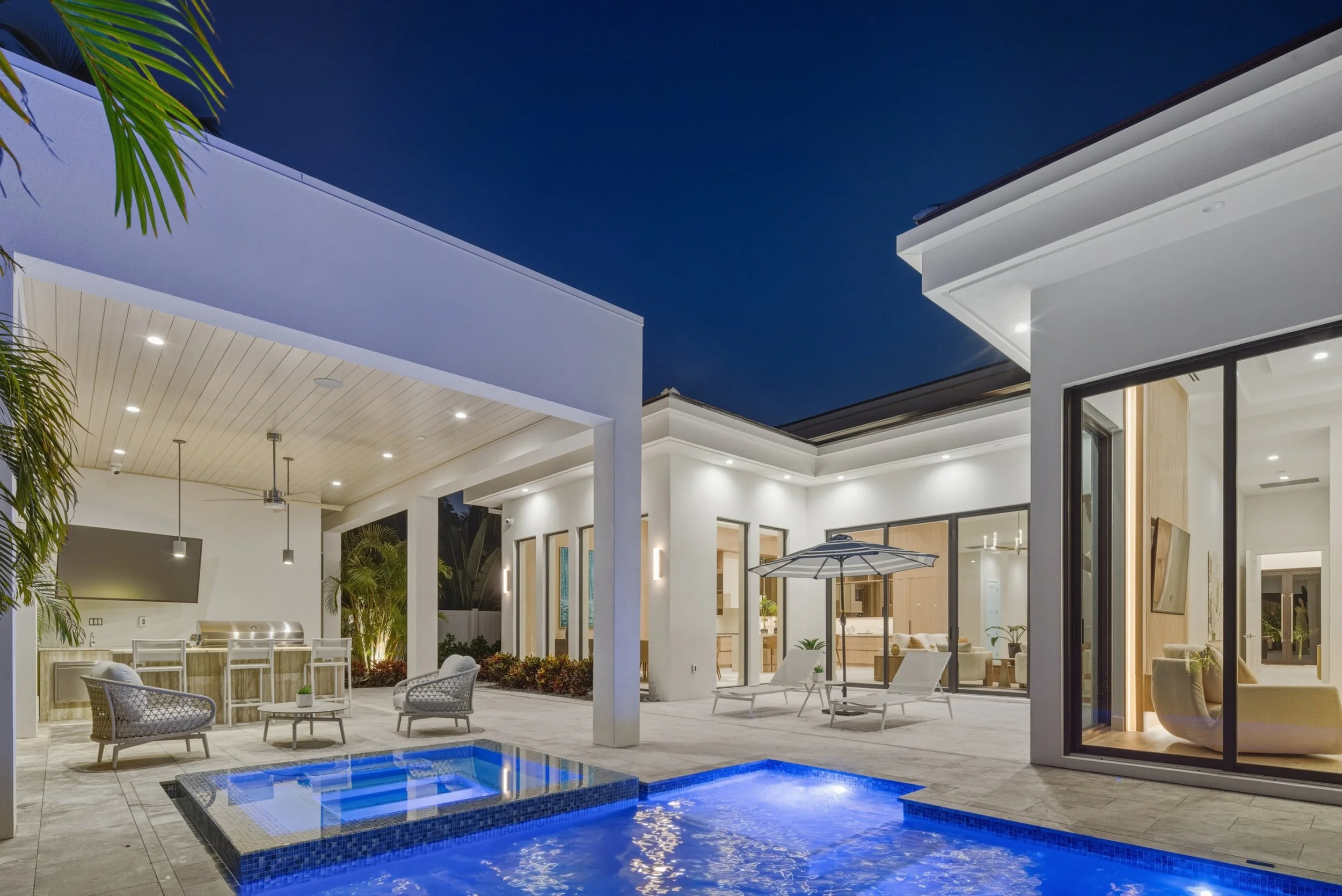 Modern house with outdoor pool area at night, illuminated with embedded lights, surrounded by patio furniture and large glass windows showing interior living space.