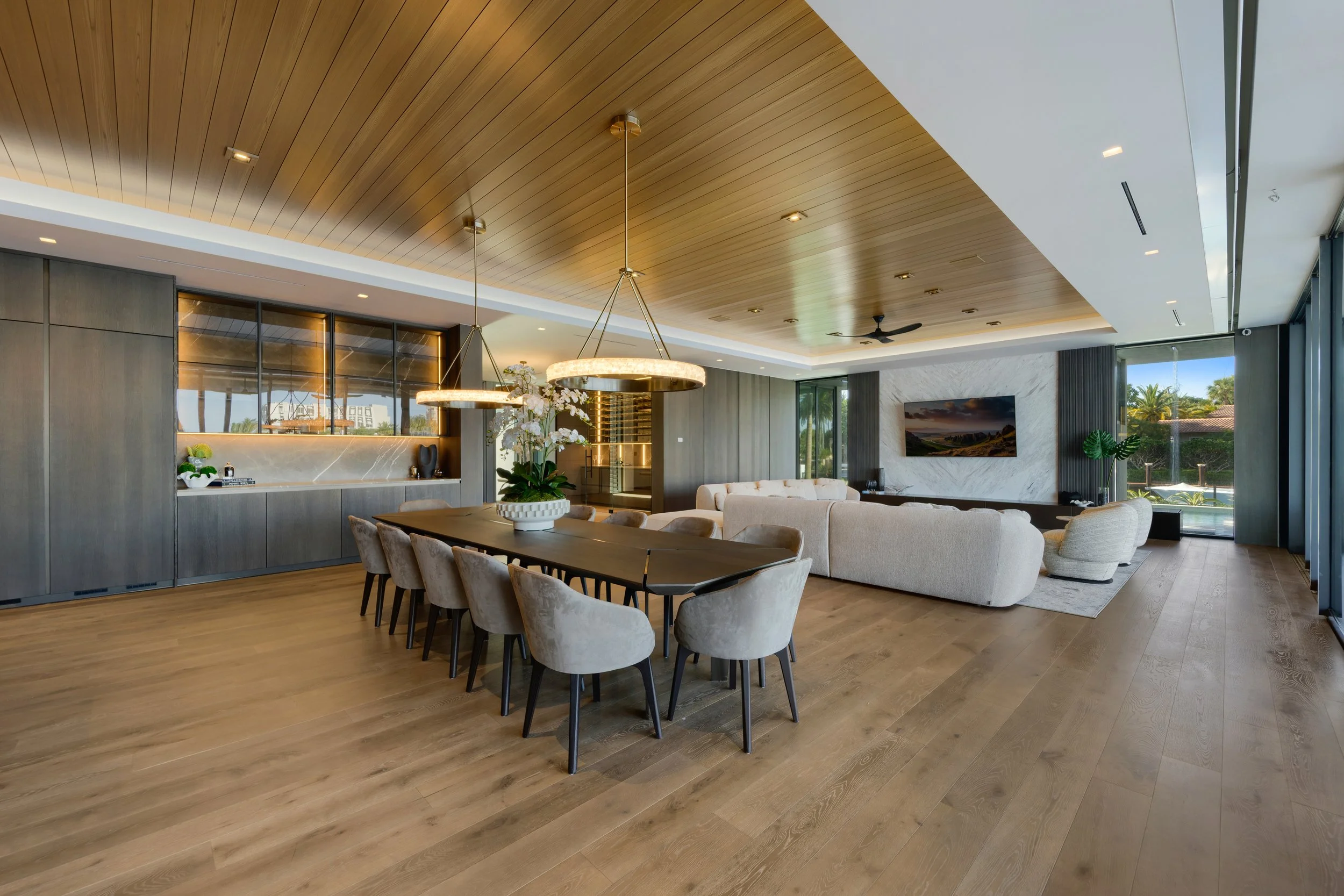 Modern living room with a wooden ceiling, large windows, white sectional sofa, dining table with beige chairs, and decorative plants.