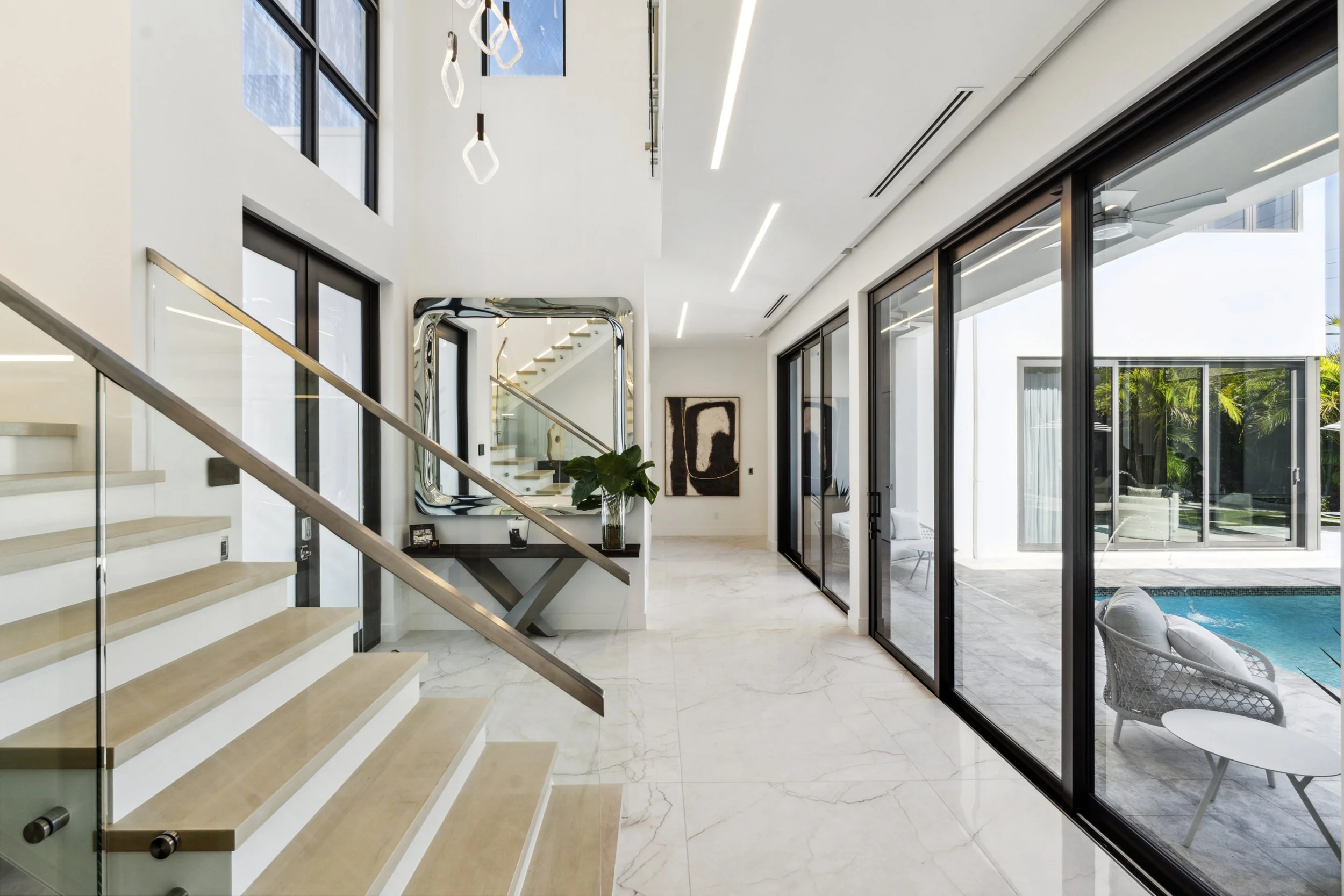 Modern interior of a house with staircase, large glass doors leading to an outdoor pool area, and minimalistic decor.
