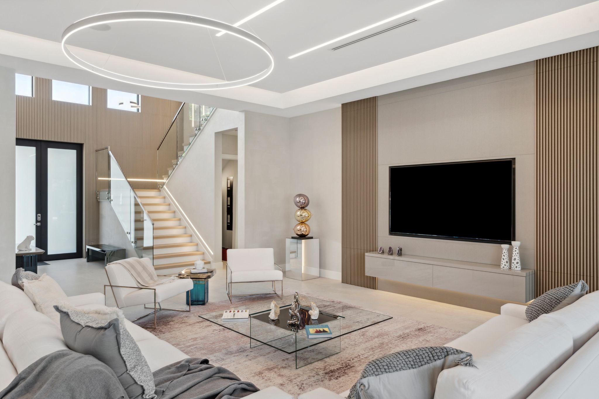 Modern living room with white sectional sofa, glass coffee table, contemporary decorations, stairs with glass railing, large flat-screen TV, and minimalist lighting fixtures.