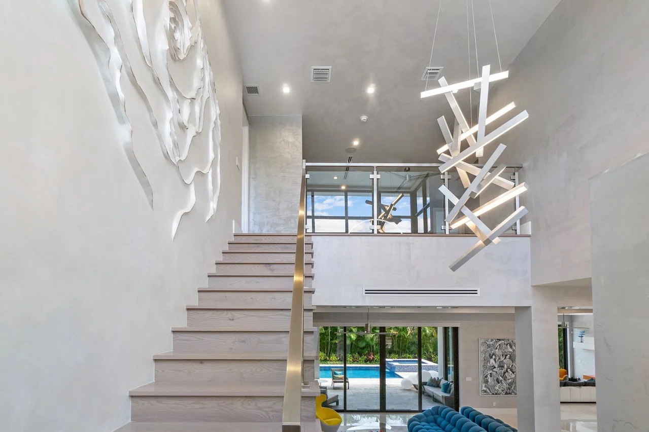 Modern interior with light wood staircase, abstract wall art, contemporary hanging light fixture, and view of outdoor pool area through glass doors.
