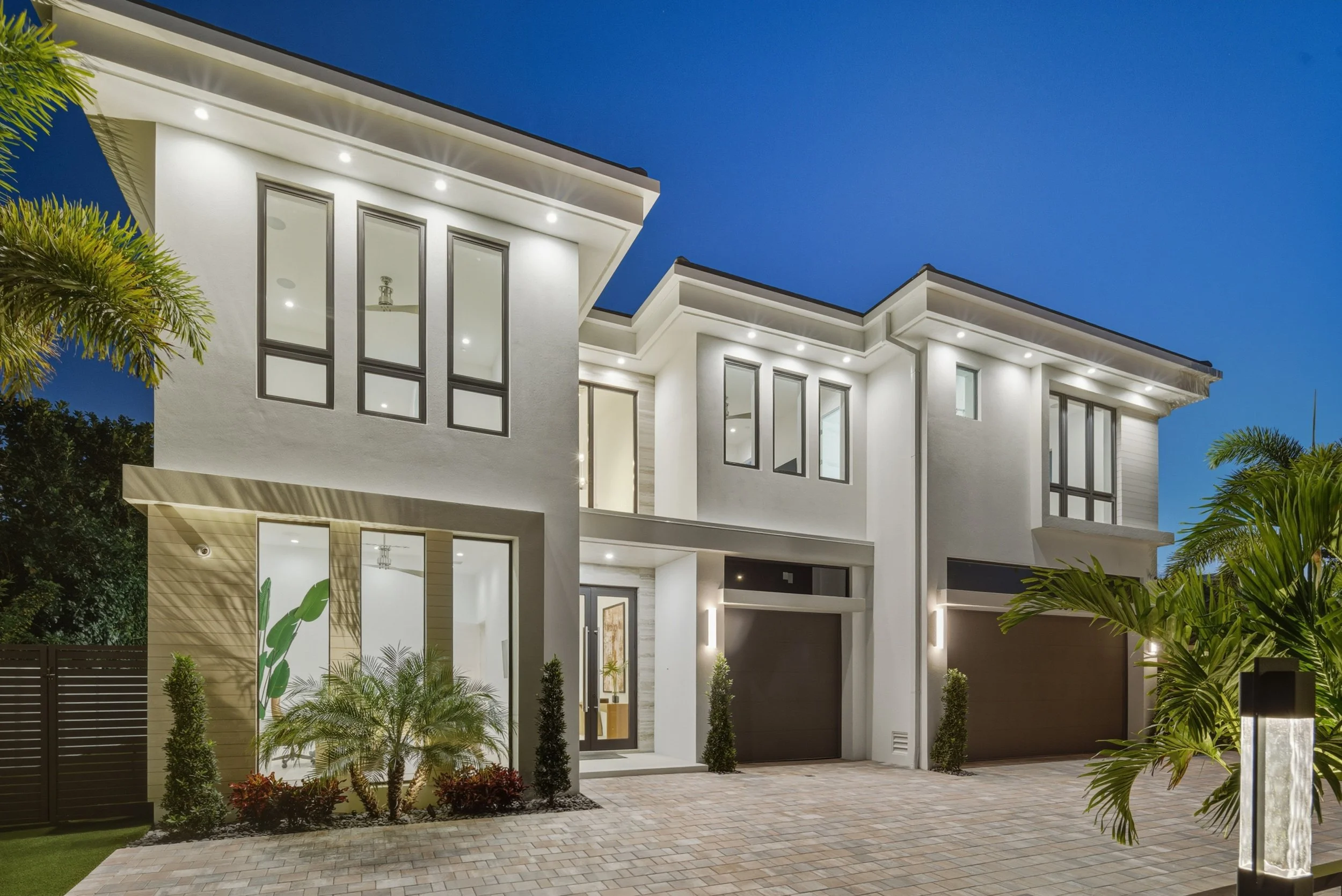 Modern white multi-story house with large windows, bright exterior lighting, surrounded by landscaped garden with small trees and plants, at dusk or nighttime.