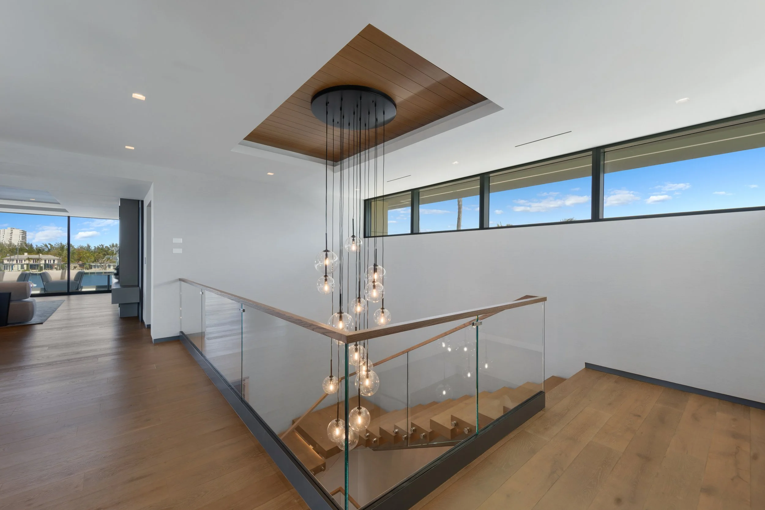 Interior of a modern, minimalist home featuring a staircase with glass railing and hanging spherical light fixtures. Large windows offer a view of the outside, showcasing a blue sky with clouds and a distant building.