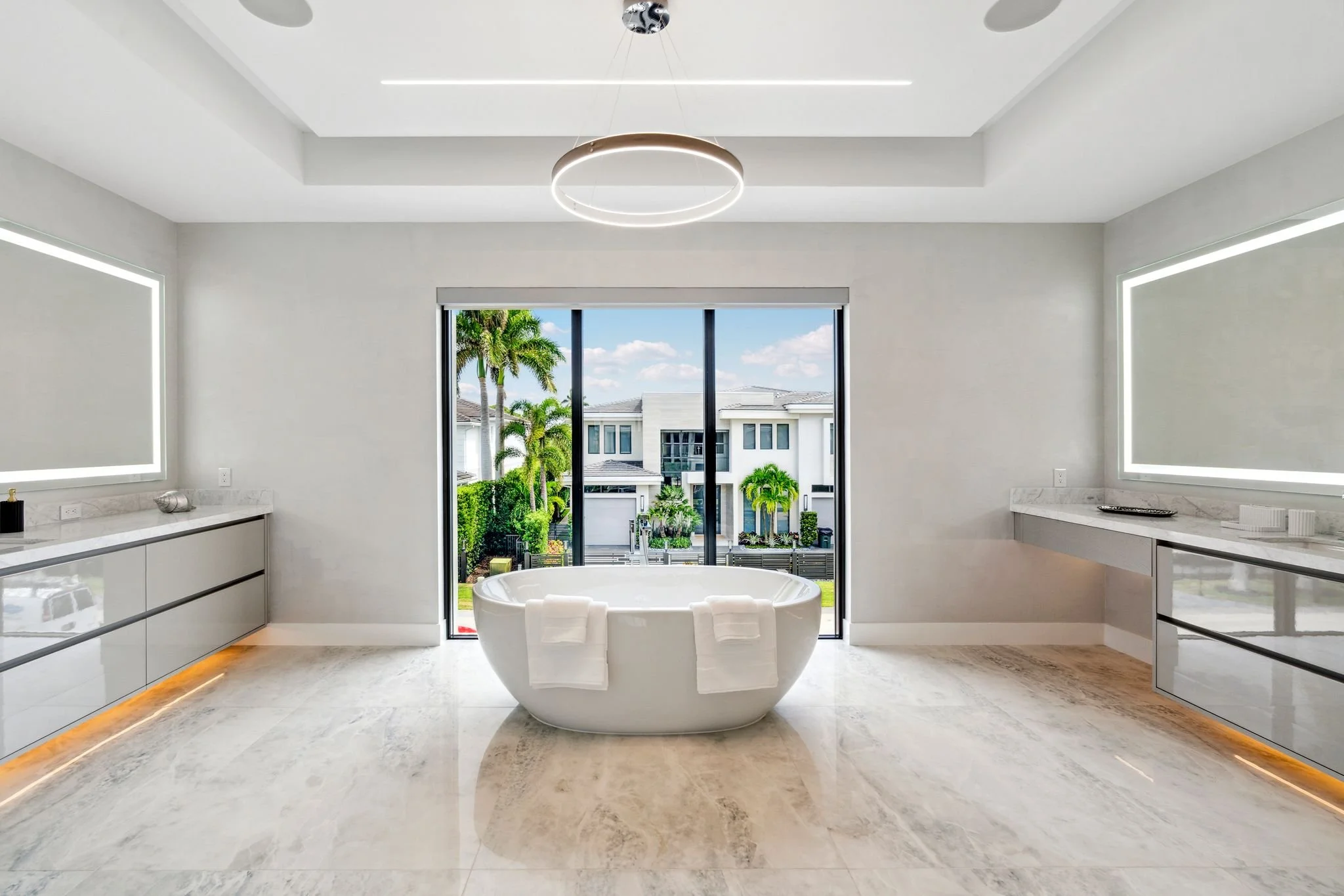 Modern bathroom with a large window showing palm trees and neighboring houses. Features include a white oval bathtub, marble countertops, and sleek, minimalistic cabinetry, with built-in LED lighting.