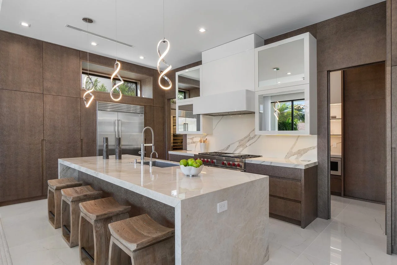 Modern kitchen with a large marble island, wooden stools, and pendant lights. White cabinets and a stainless steel stove are visible. Windows let in natural light, and a bowl of green apples is on the island.