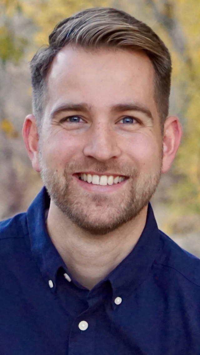 A smiling man with short light brown hair and blue eyes, wearing a navy blue button-up shirt, outdoors with blurred autumn trees in the background.