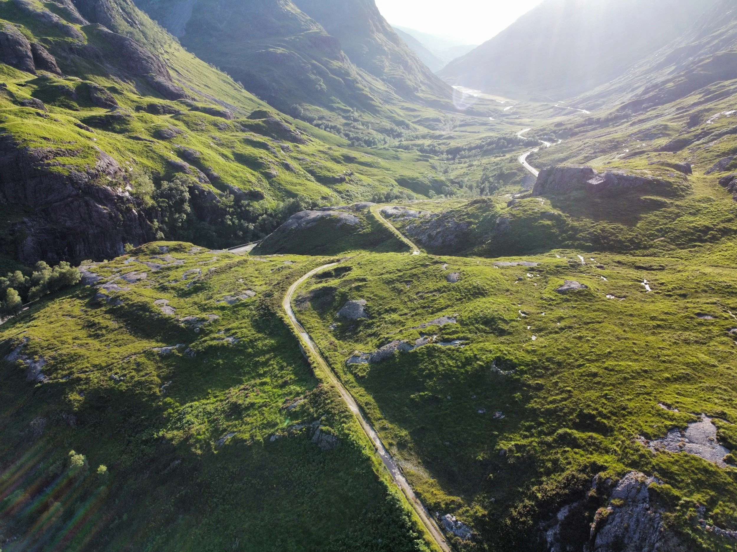Aerial view of a lush green mountainous landscape with winding roads through the valley, and sunlight illuminating the scene.
