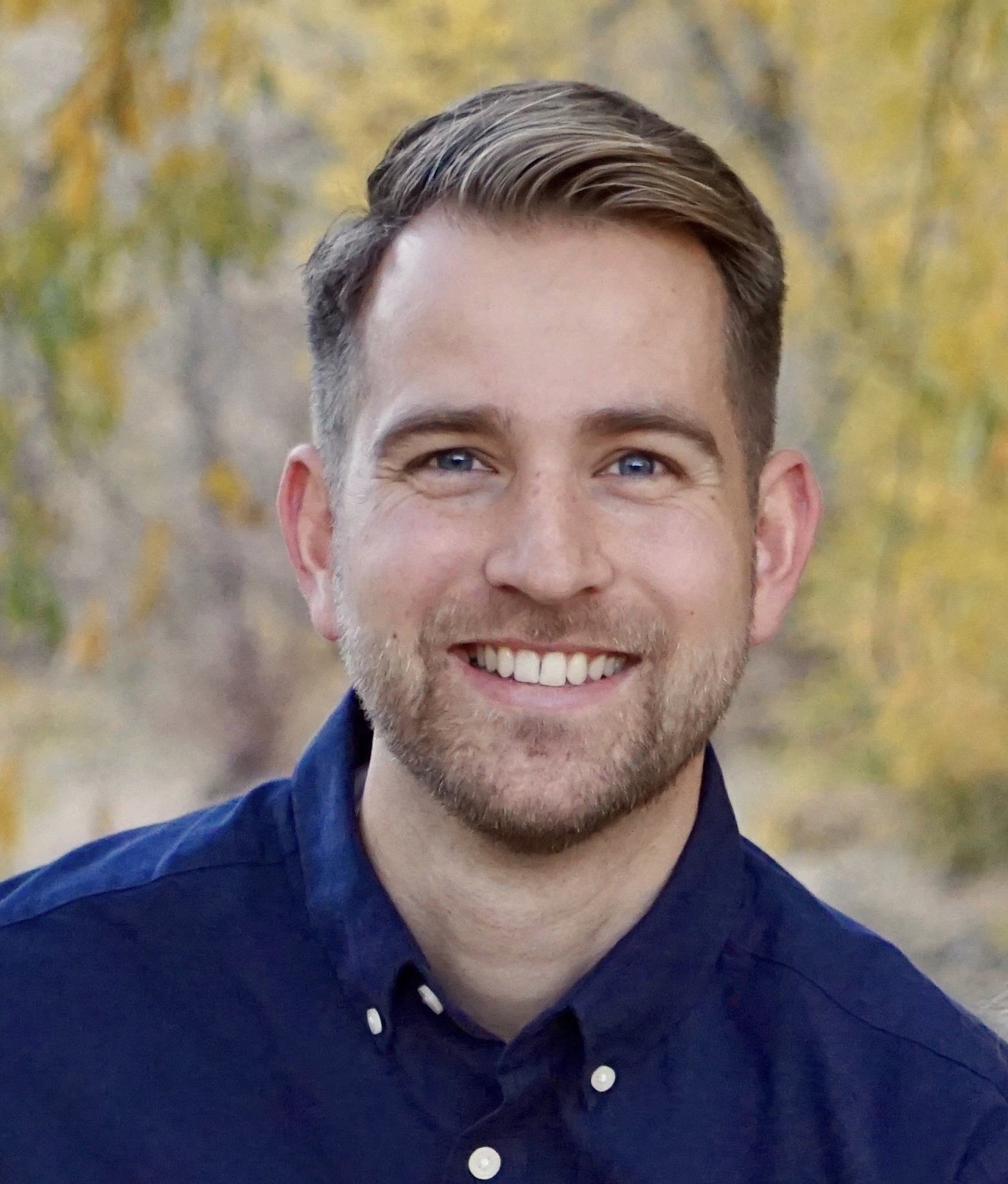 A man with short brown hair, blue eyes, and stubble, smiling outdoors with autumn trees in the background. He is wearing a dark blue button-up shirt.