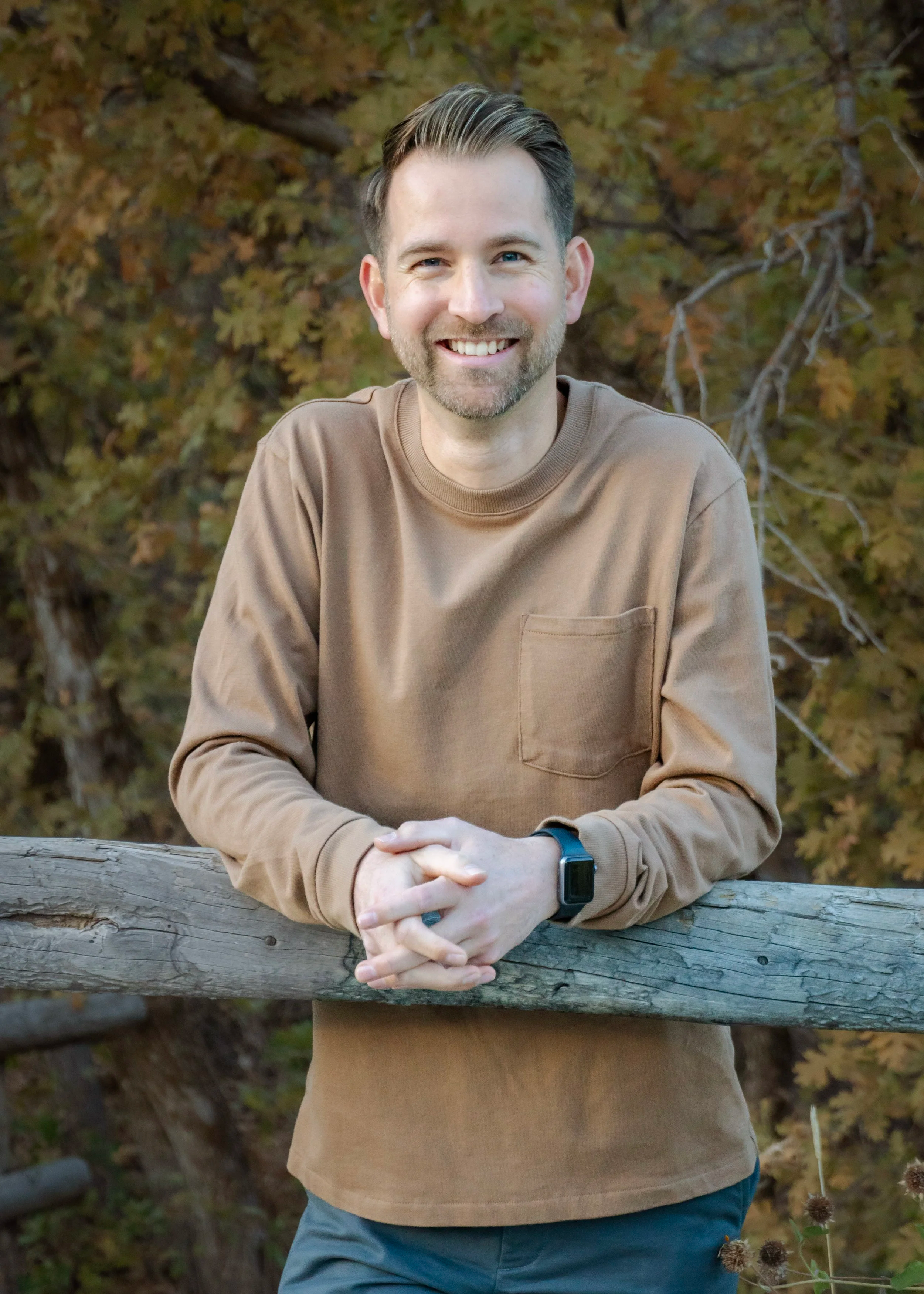 A smiling man with short, dark hair, a beard, and a smartwatch on his left wrist, leaning on a wooden railing outdoors during autumn with yellow and brown leaves in the background.