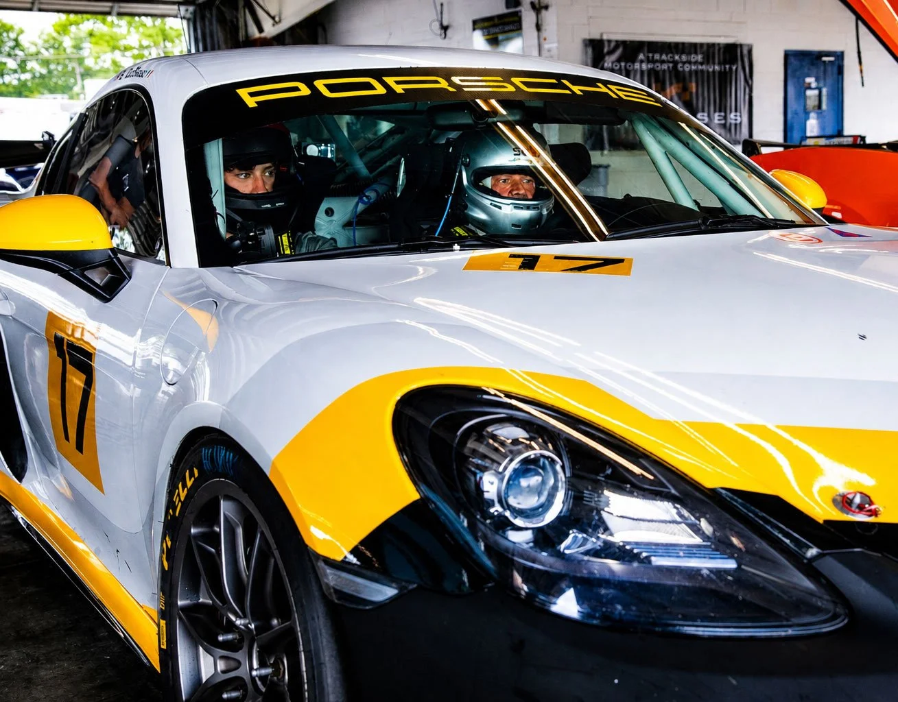 Two race car drivers in helmets sitting in a white and yellow Porsche race car inside a garage.