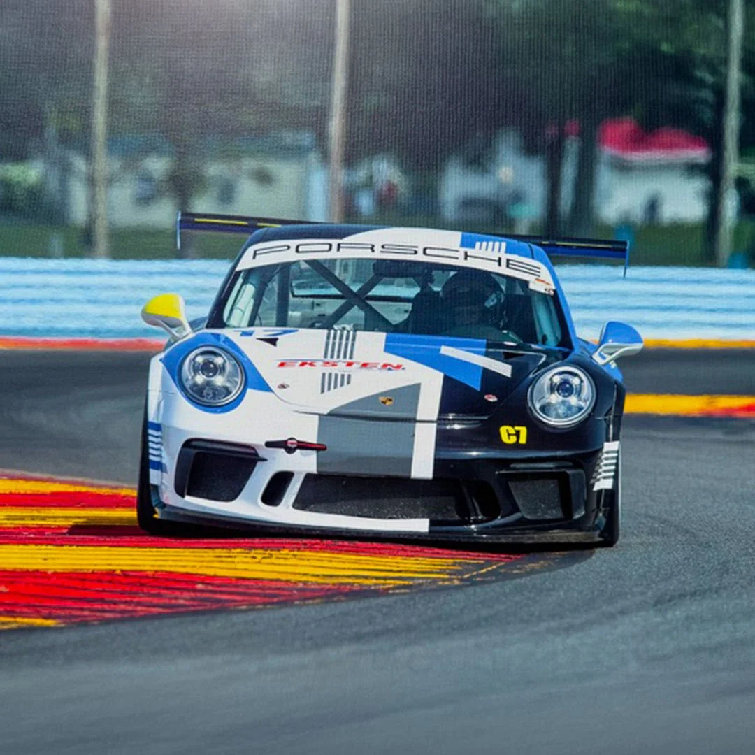 A Porsche race car on a racetrack during a race, with a blue, white, and black paint job and yellow side mirror and C7 label.