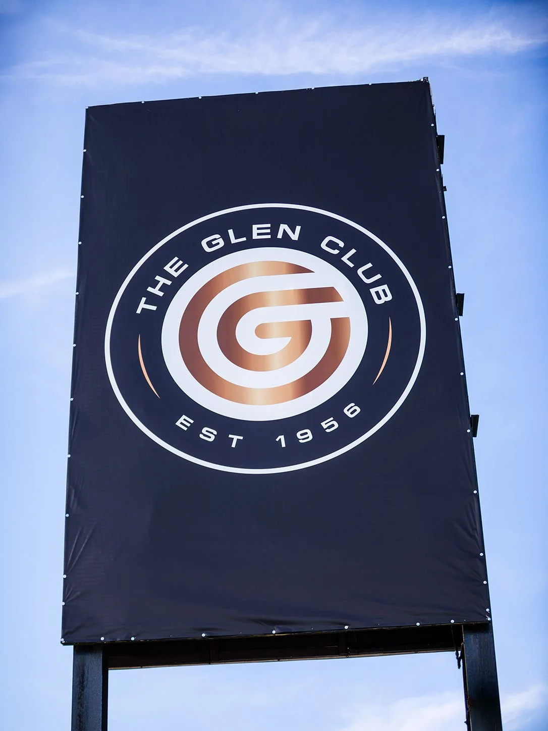Large black banner with a circular logo for The Glen Club, established 1956, featuring a stylized 'G' in the center, mounted on a stand against a blue sky with few clouds.