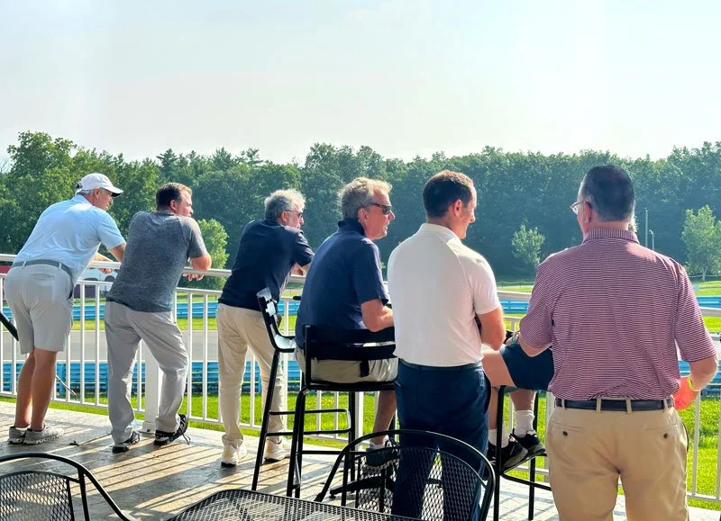 Group of six men standing and sitting on the balcony of a golf course, looking out at the green landscape and the race track.