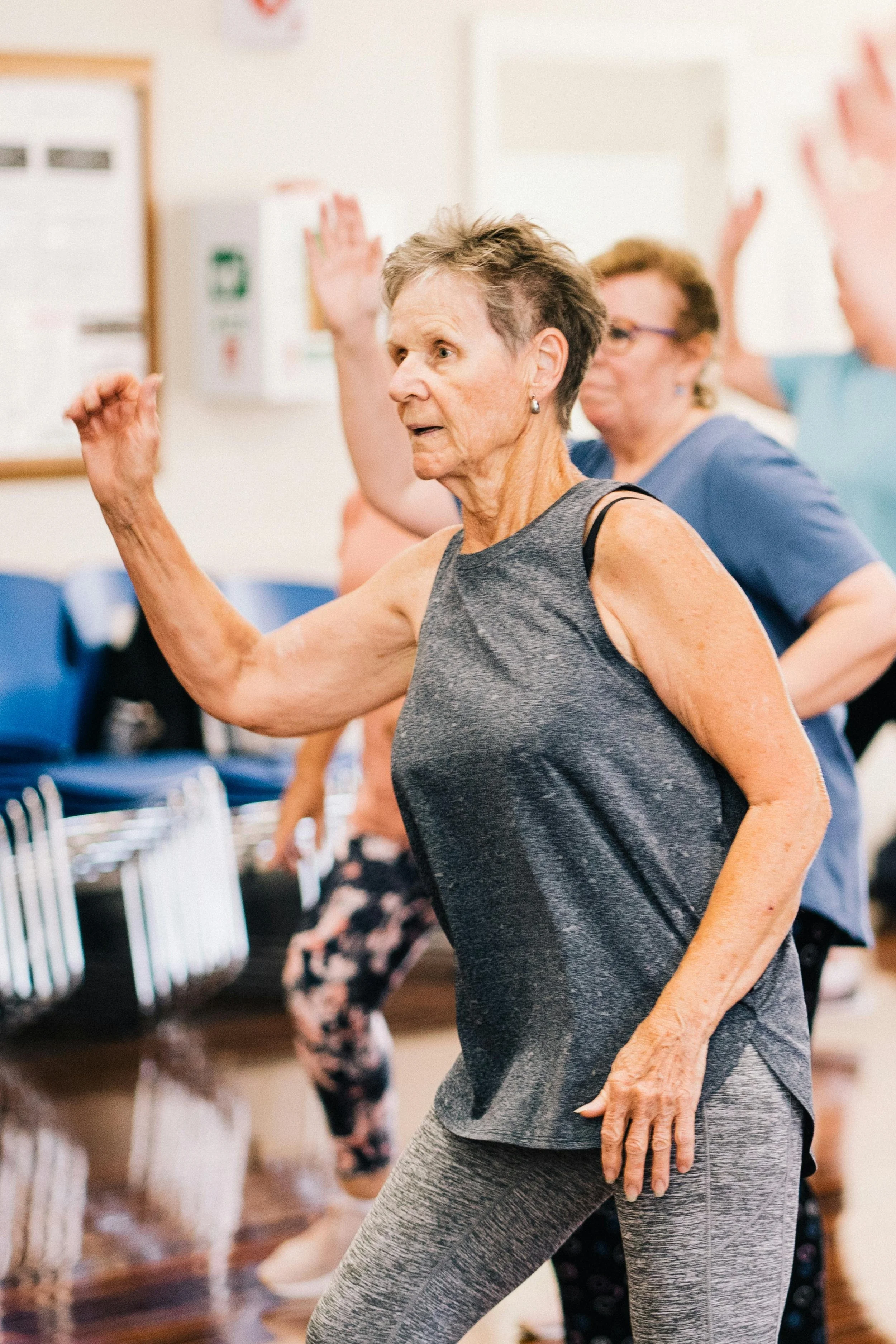Older woman participating in a group exercise class, raising her right arm and moving to music.