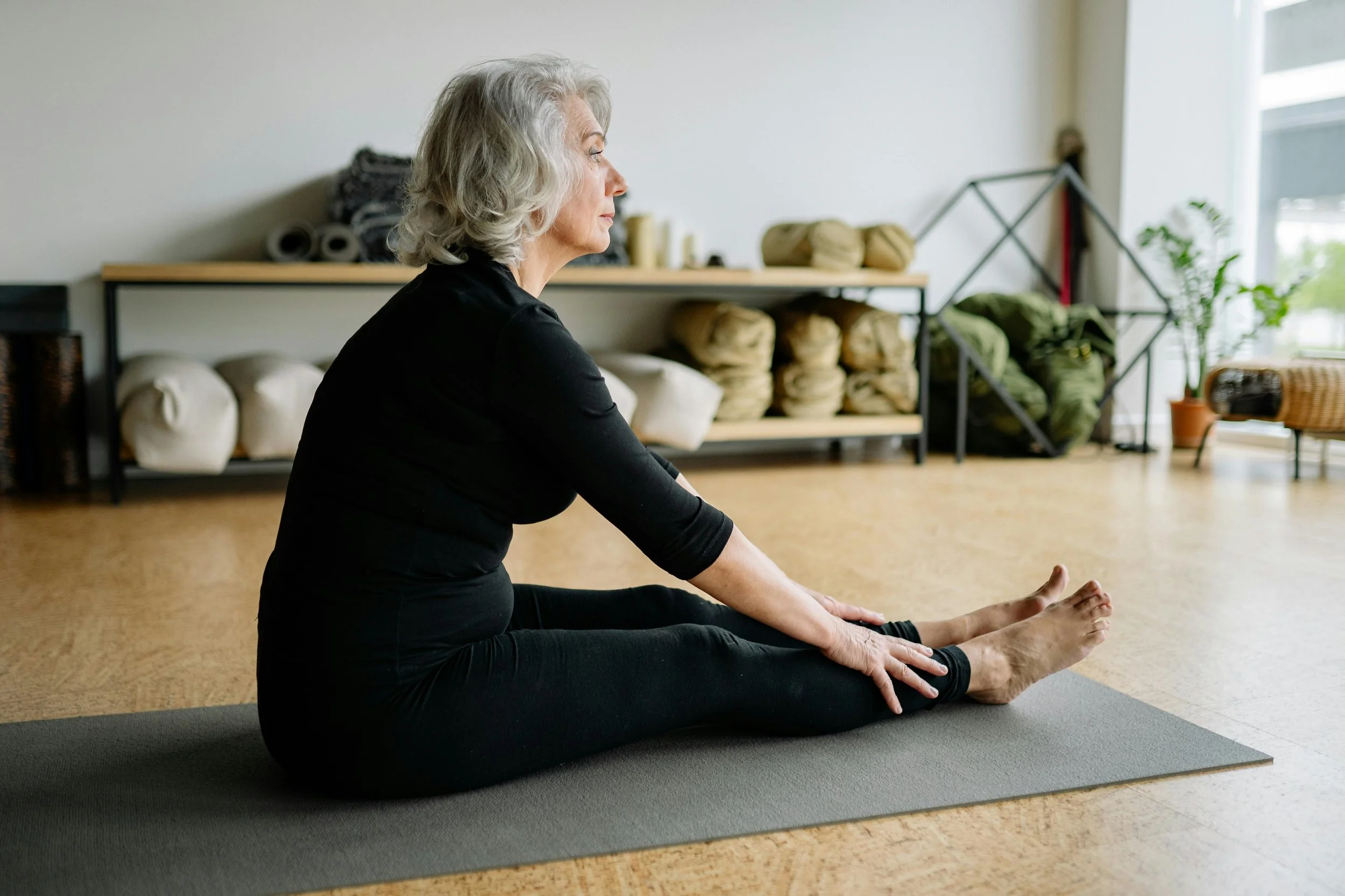An elderly woman with gray hair practicing yoga in a studio, sitting with legs extended and hands on her feet, with yoga mats and supplies stored on shelves in the background.