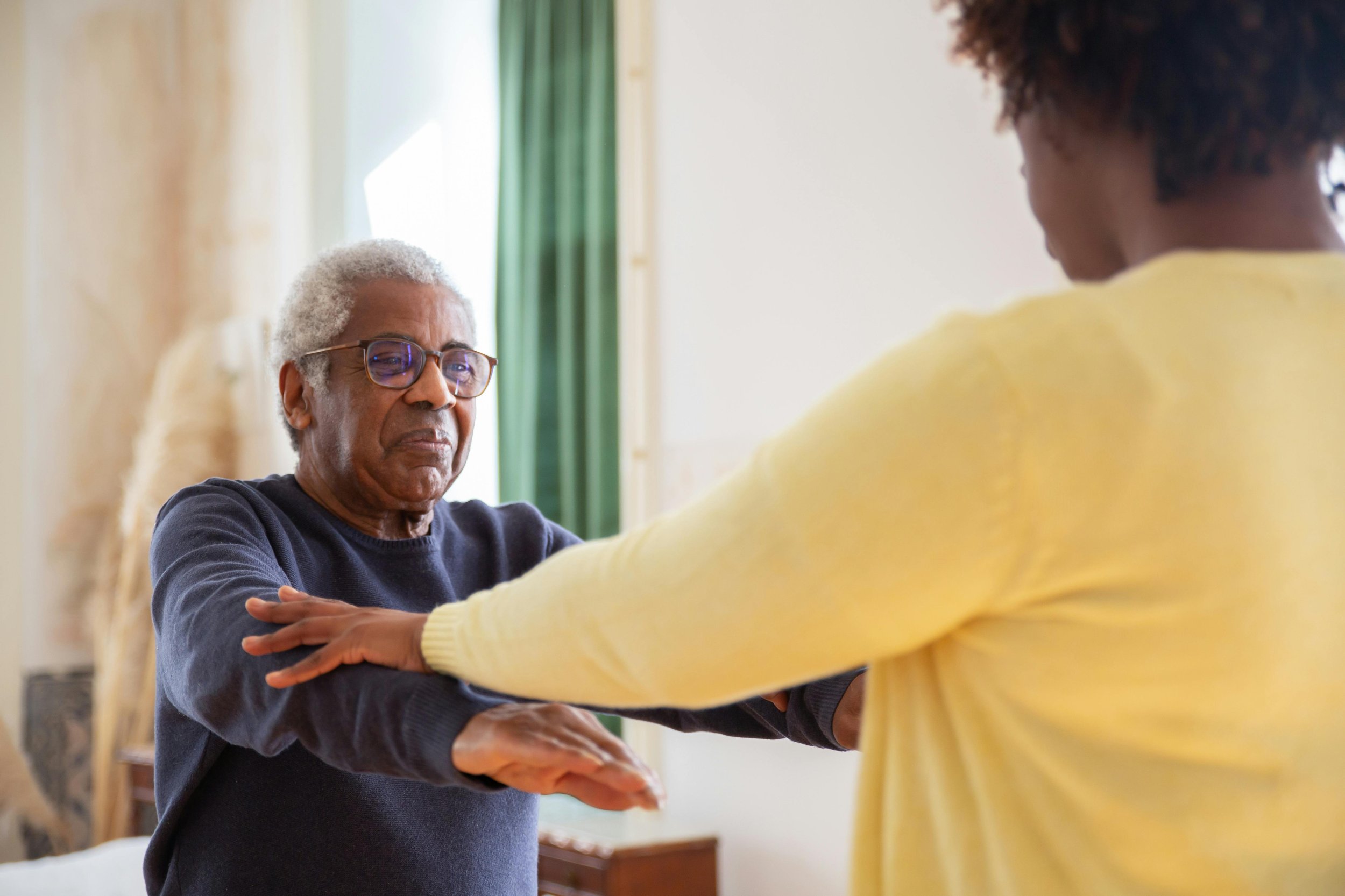 An elderly man with gray hair and glasses is holding his arms out in front of a woman in a yellow sweater. The woman is standing close to him, touching his arms, with her back to the camera. They appear to be engaged in a therapy or rehabilitation activity in a well-lit room.