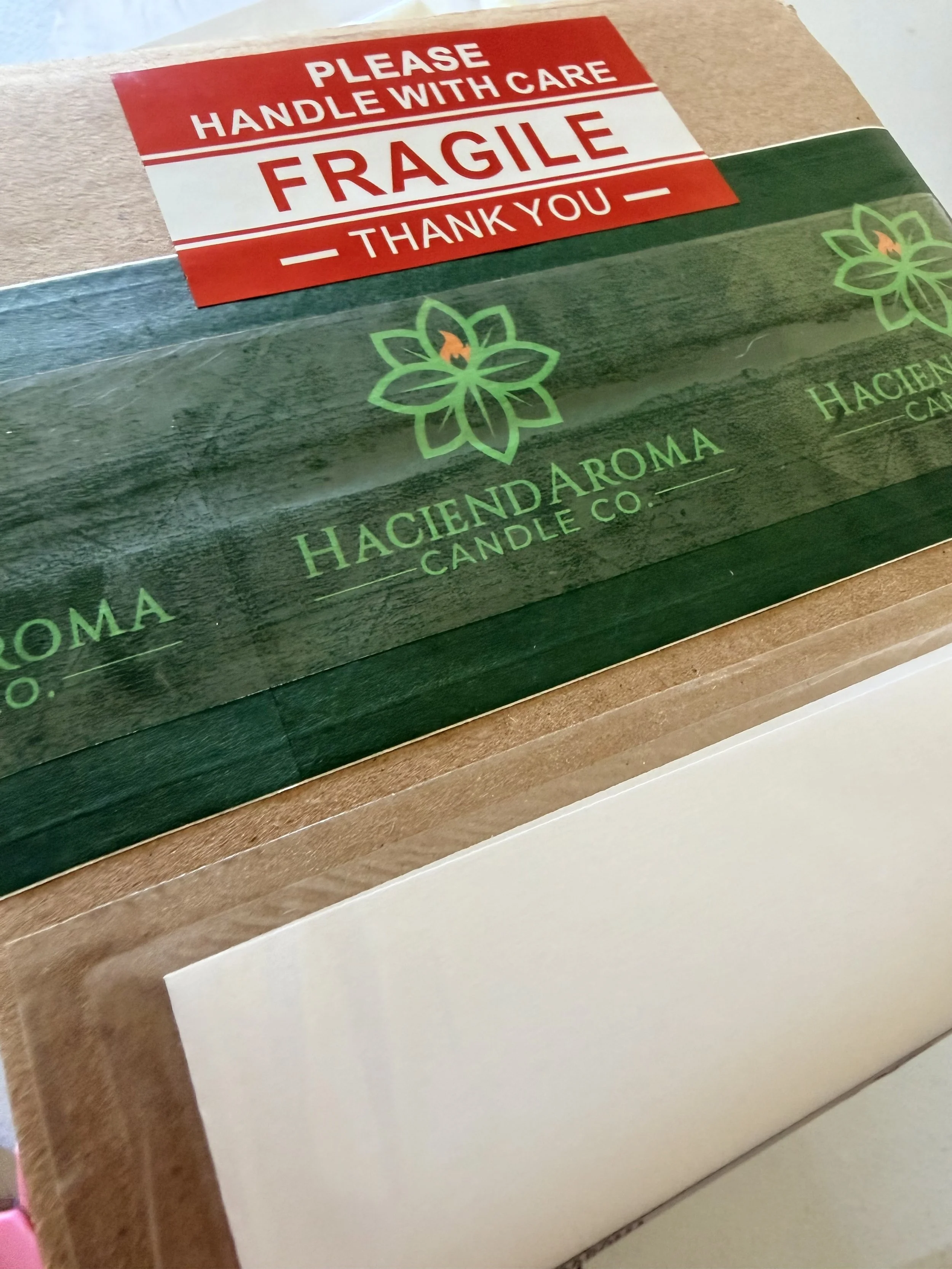 Close-up of a package with a red and white label reading 'Please Handle With Care, Fragile, Thank You' and a green logo of a flower or leaf with a flame in the center. The package appears to be made of cardboard.