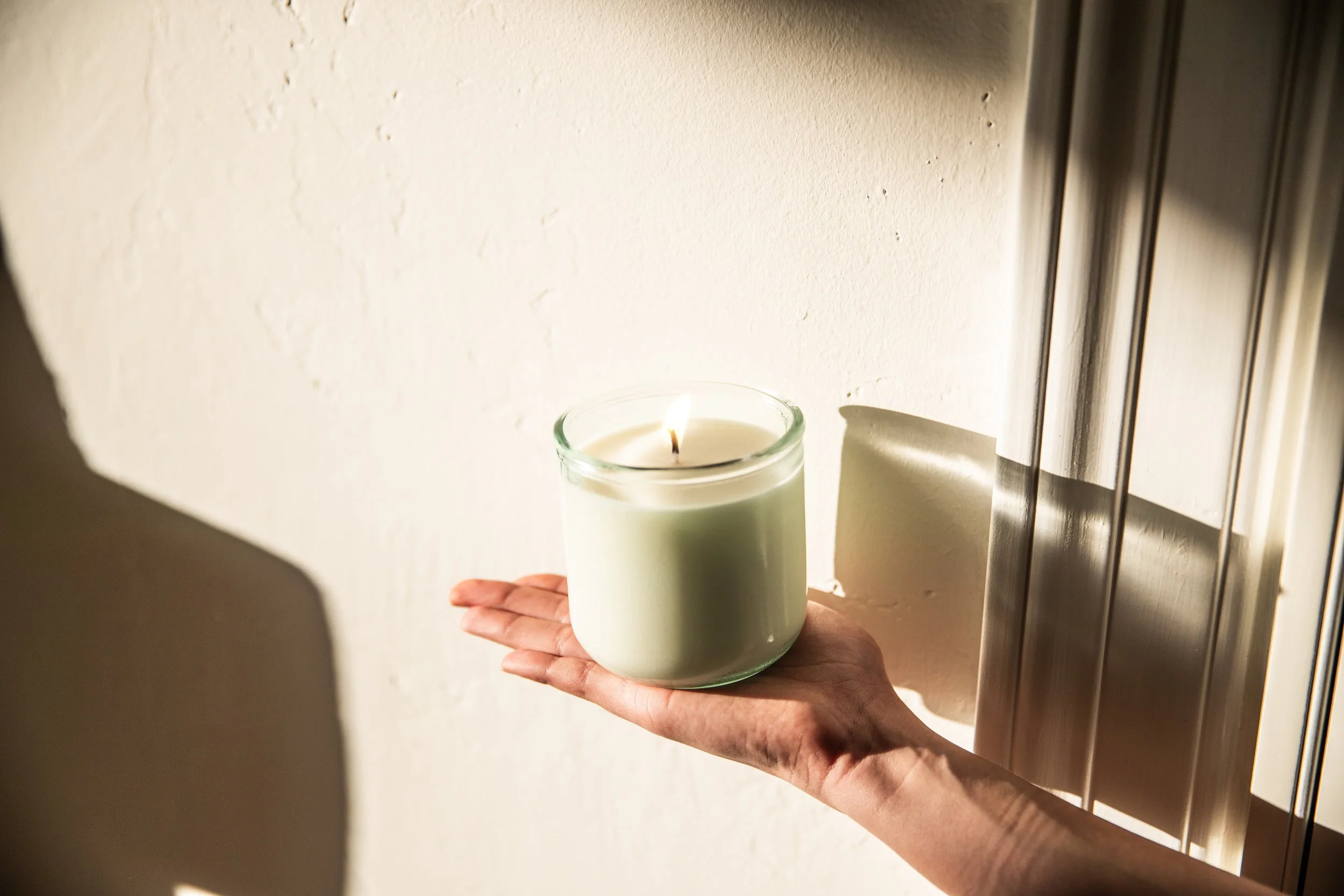A hand holding a glass candle holder with a lit white candle on a countertop near a wall and a refrigerator.
