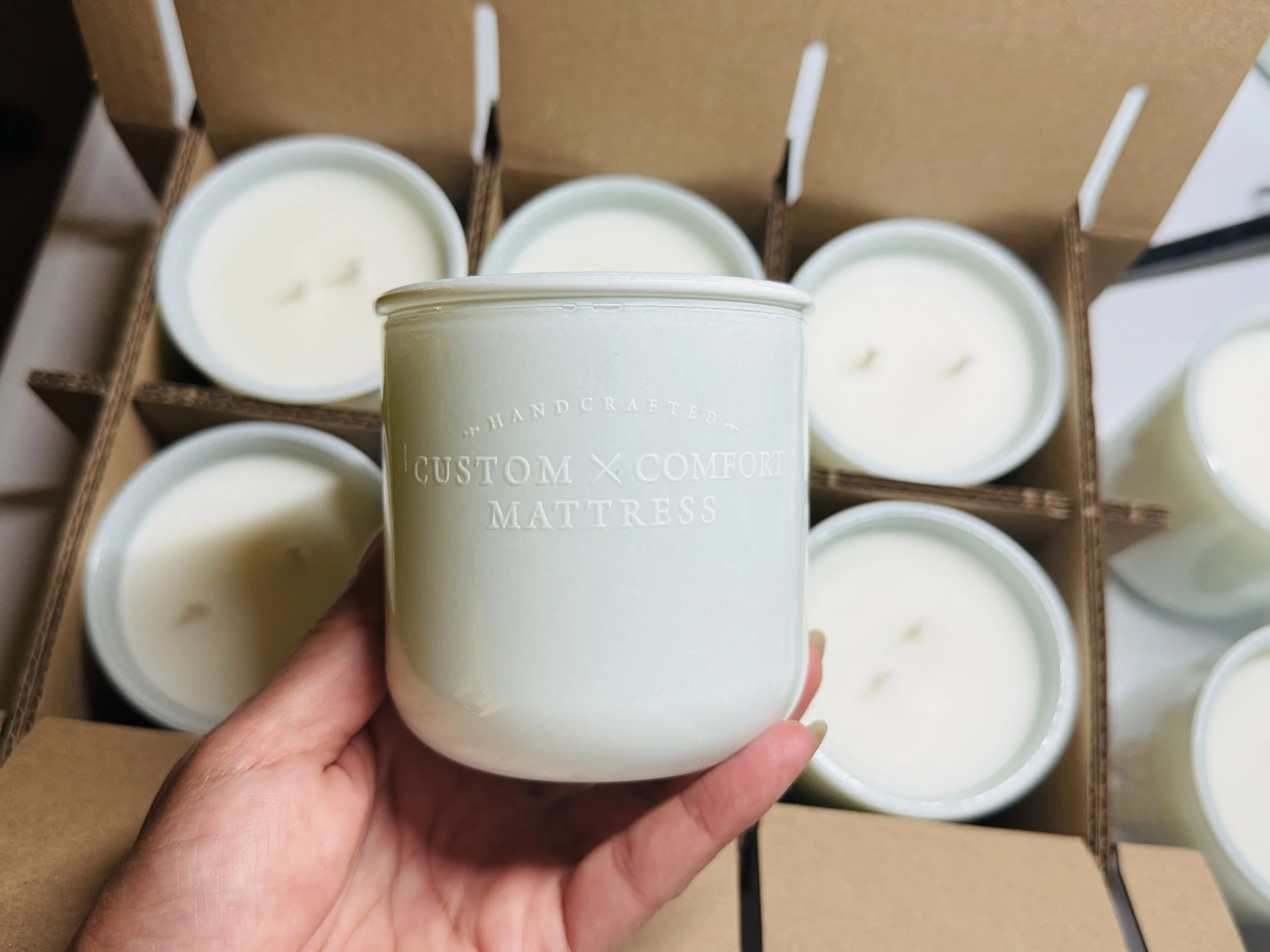 Hand holding a white candle with embossed text in front of open cardboard boxes filled with similar candles.