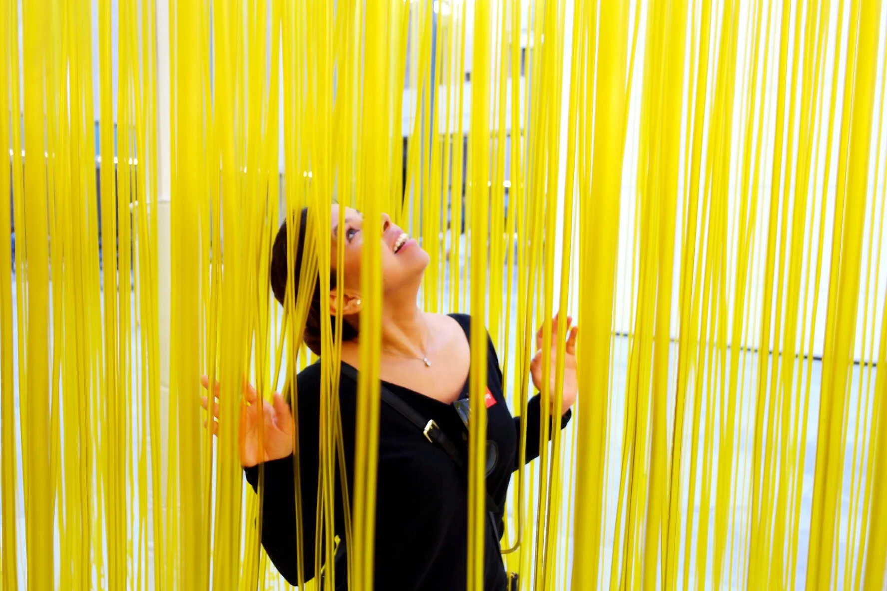 Woman with short dark hair and black top looking upward, standing behind many yellow vertical ribbons or streams.