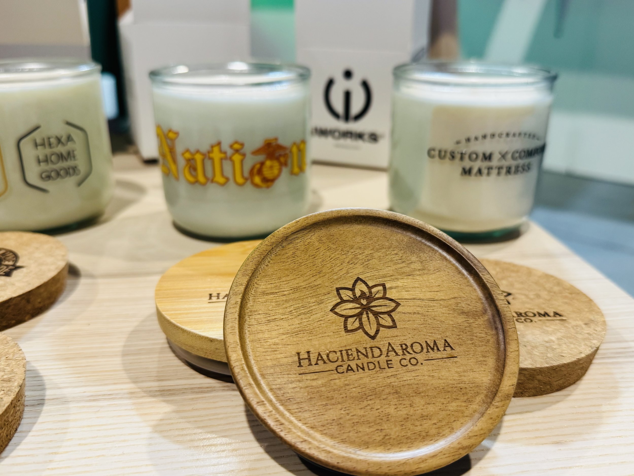 Close-up of wooden candle lid with engraved logo from Hacienda Aroma Candle Co., and various candles in glass containers in the background.