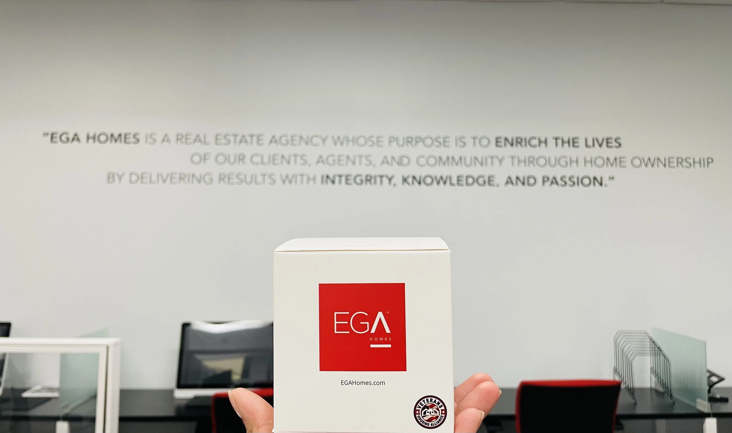 A hand holding a white and red sign that says "EGA HOMES" in front of a conference room background with chairs, table, and a sign on the wall outlining the company's purpose.