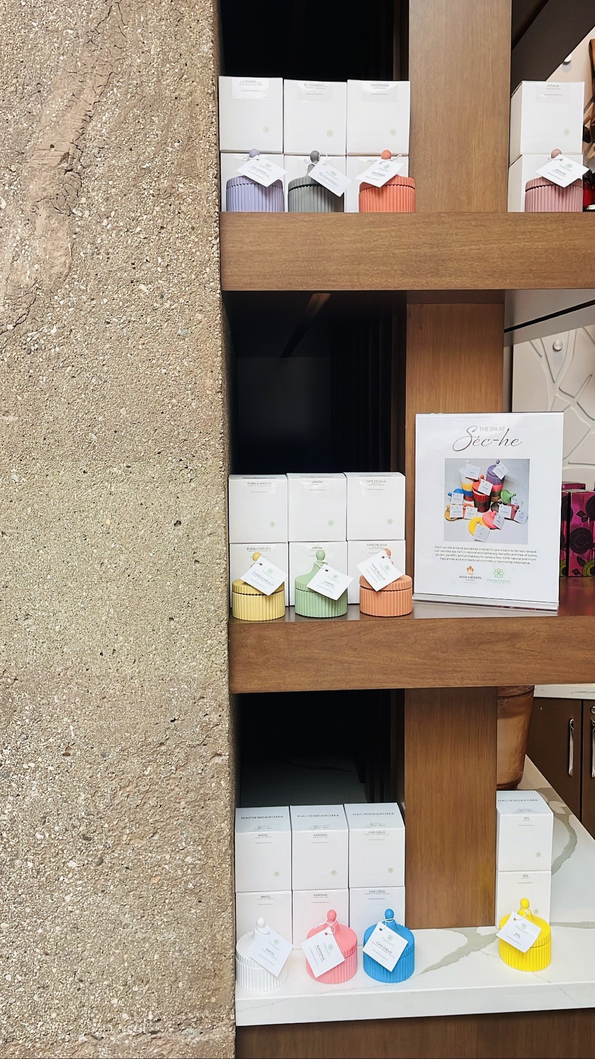 Colorful wax candles in various shapes and sizes displayed on wooden shelves, with packaging boxes and a printed sign nearby.