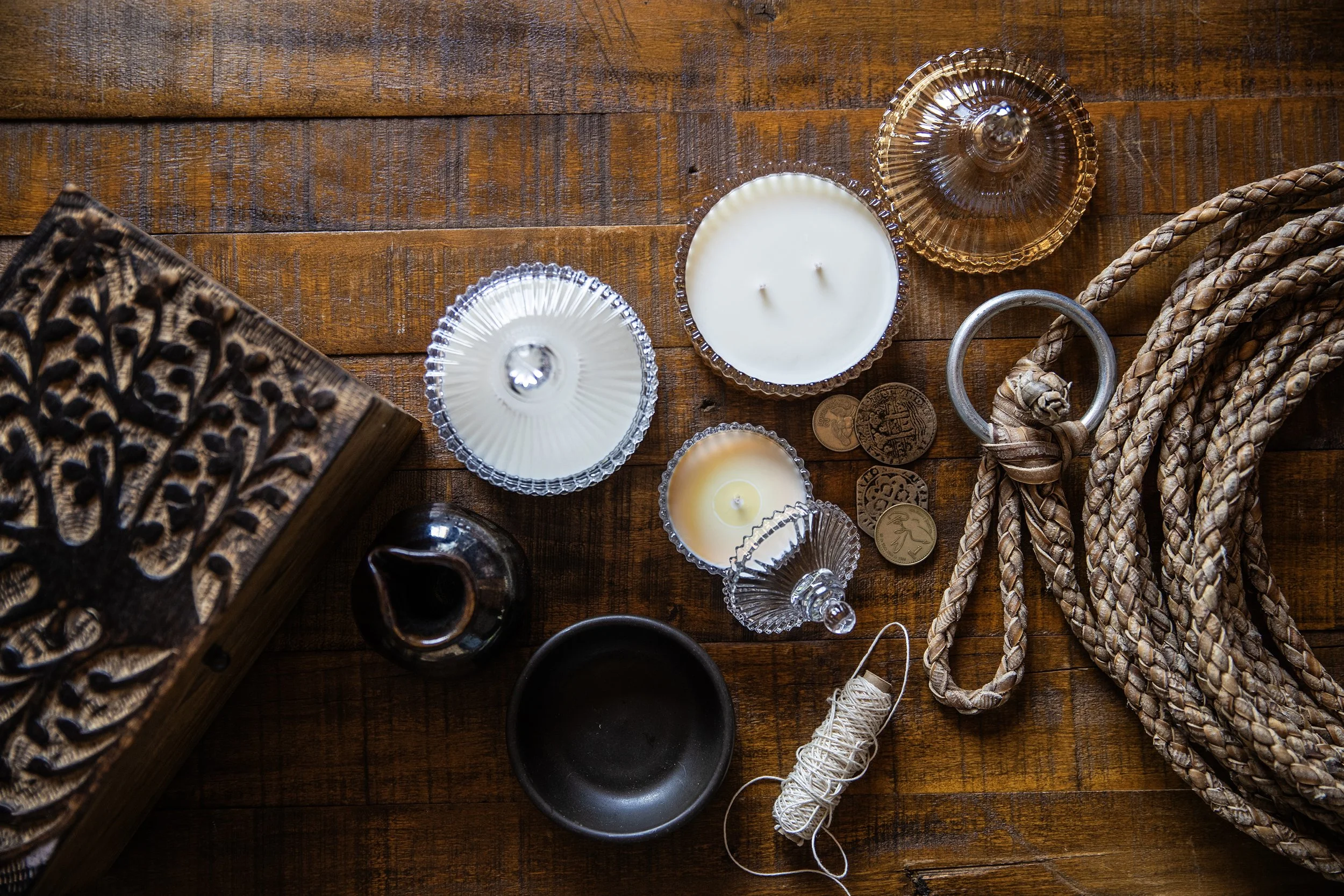 Top-down view of a wooden table with candles in glass holders, decorative coins, a woven rope, and a carved wooden box.