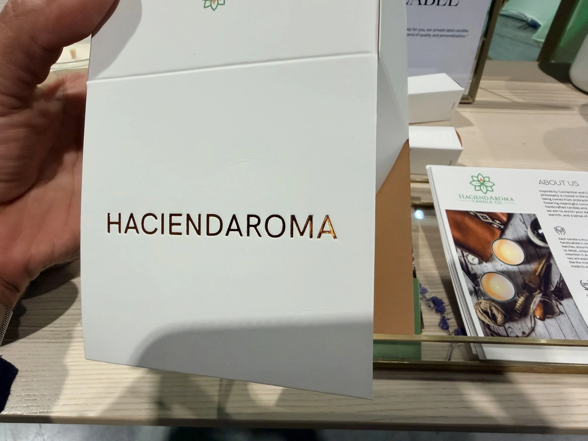 Close-up of a white box labeled 'HACIENDA' being held by a person's hand, with other informational booklets or brochures on a wooden table in the background.