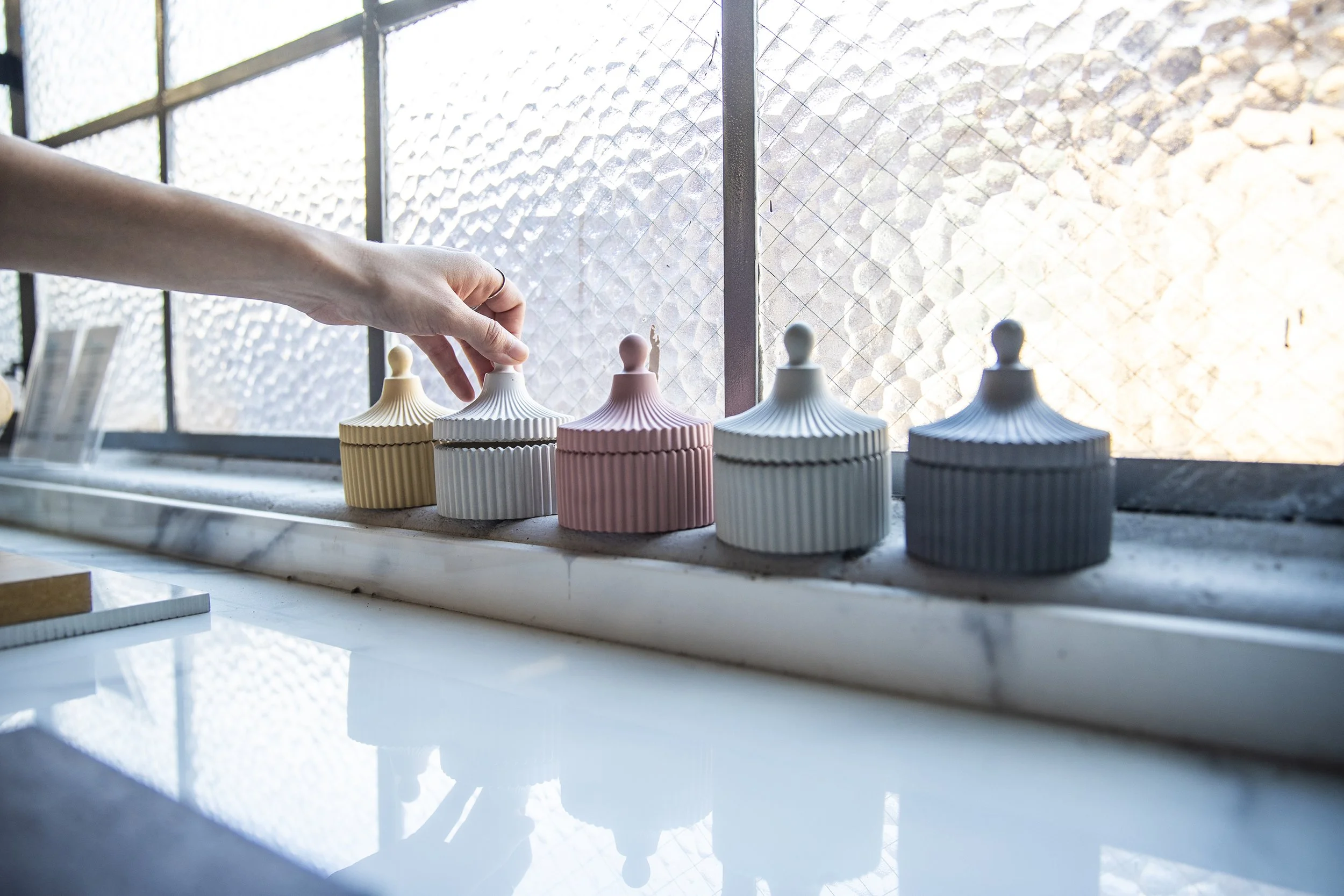 A person's hand reaches out to pick up a ceramic container with a lid, among other similar containers in pastel colors, placed on a windowsill with natural sunlight.