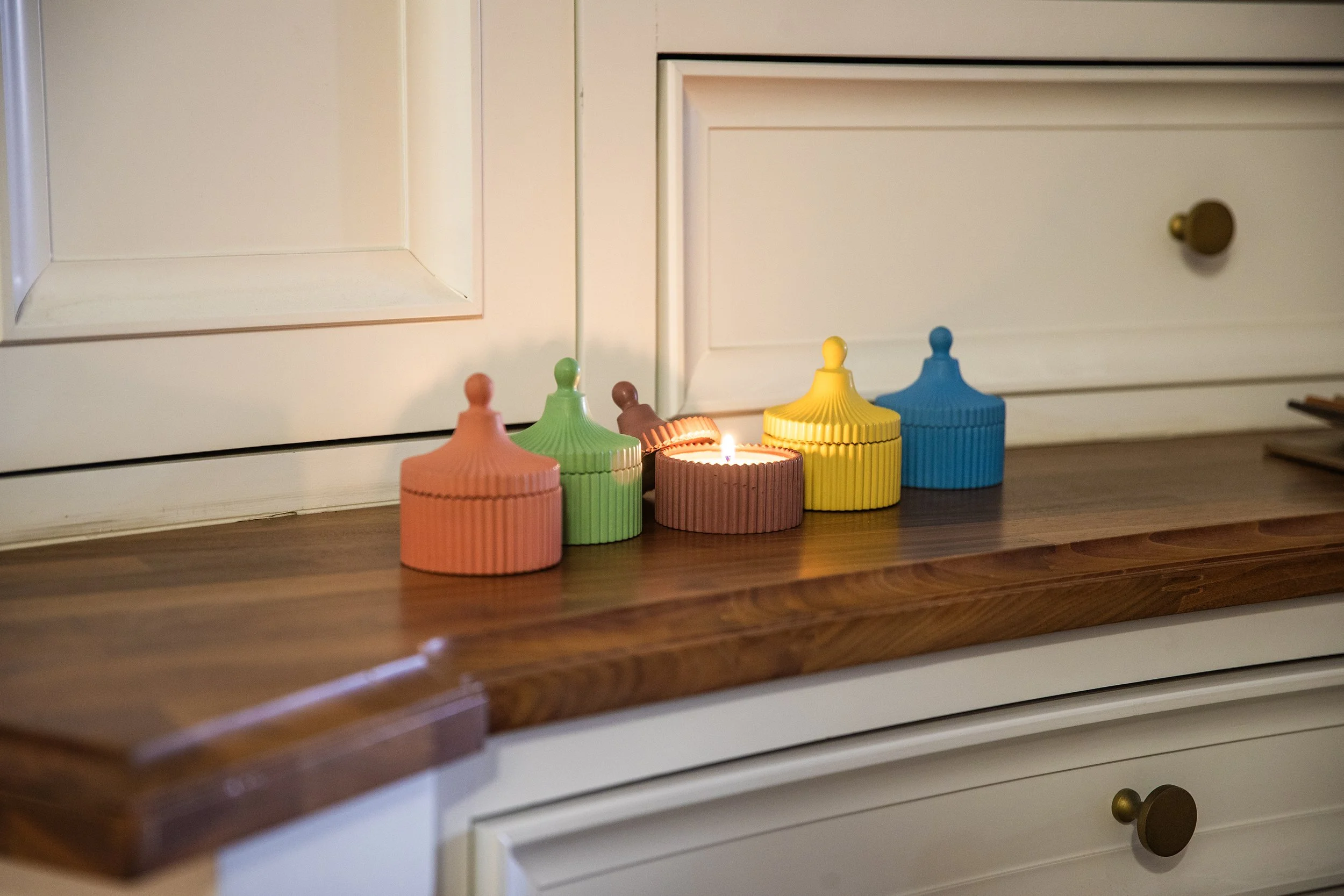 A row of colorful candles with lids placed on a wooden surface in front of white cabinets.
