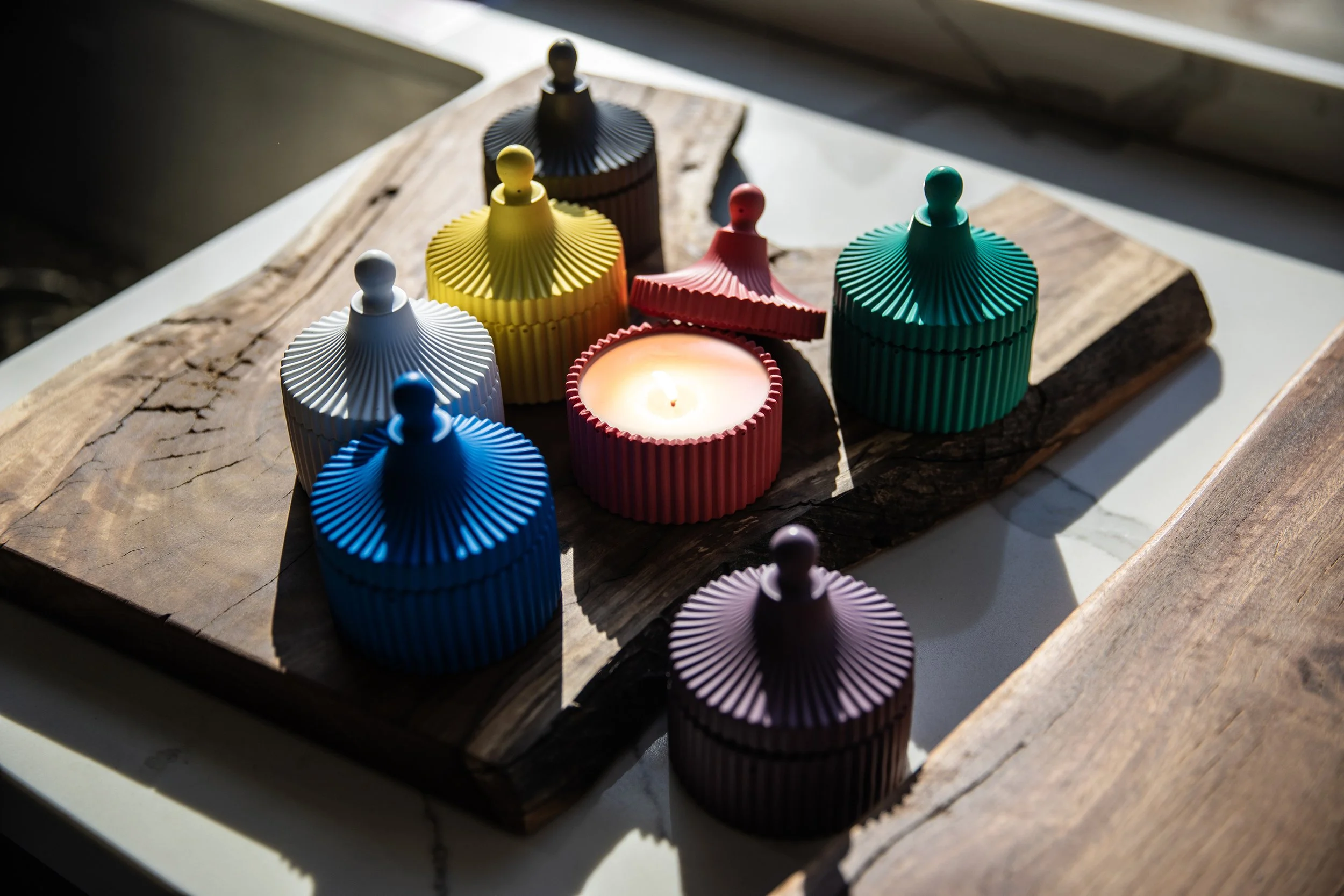 Colorful candles with ribbed texture in various colors placed on a wooden surface, with one lit candle.