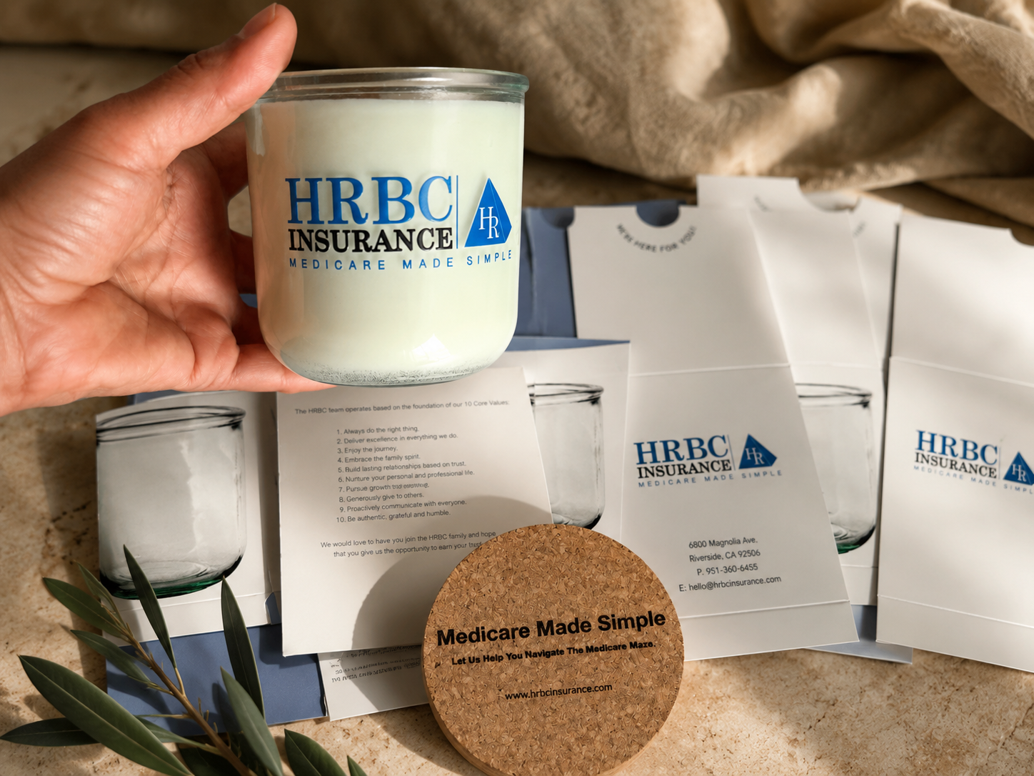 A hand holding a candle with the HRBC Insurance logo, surrounded by HRBC Insurance informational flyers and a round cork coaster with the text "Medicare Made Simple" on a beige surface with a small branch of greenery.