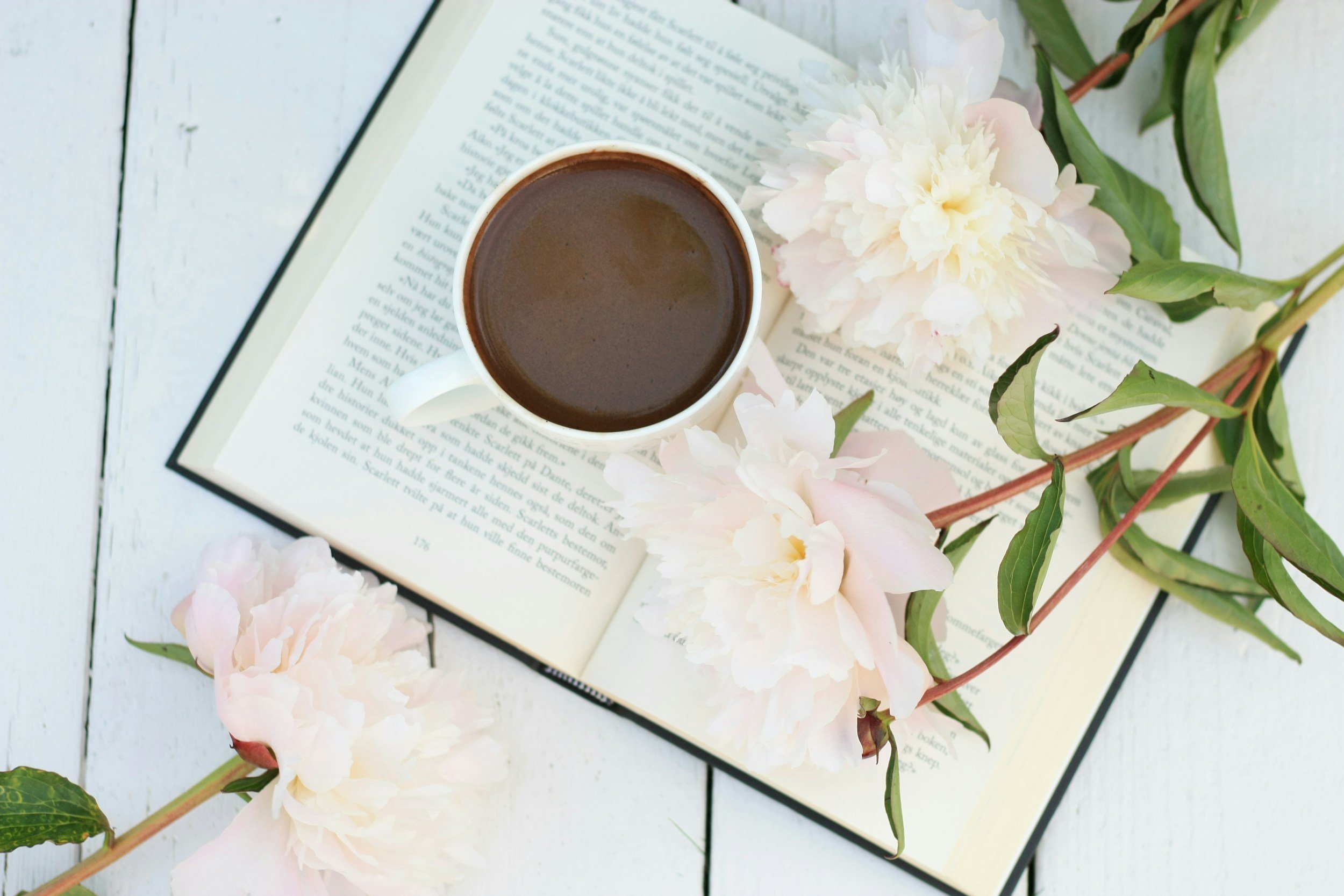 A cup of hot coffee on top of an open book, surrounded by pink and white flowers, and green leaves on a white wooden surface.
