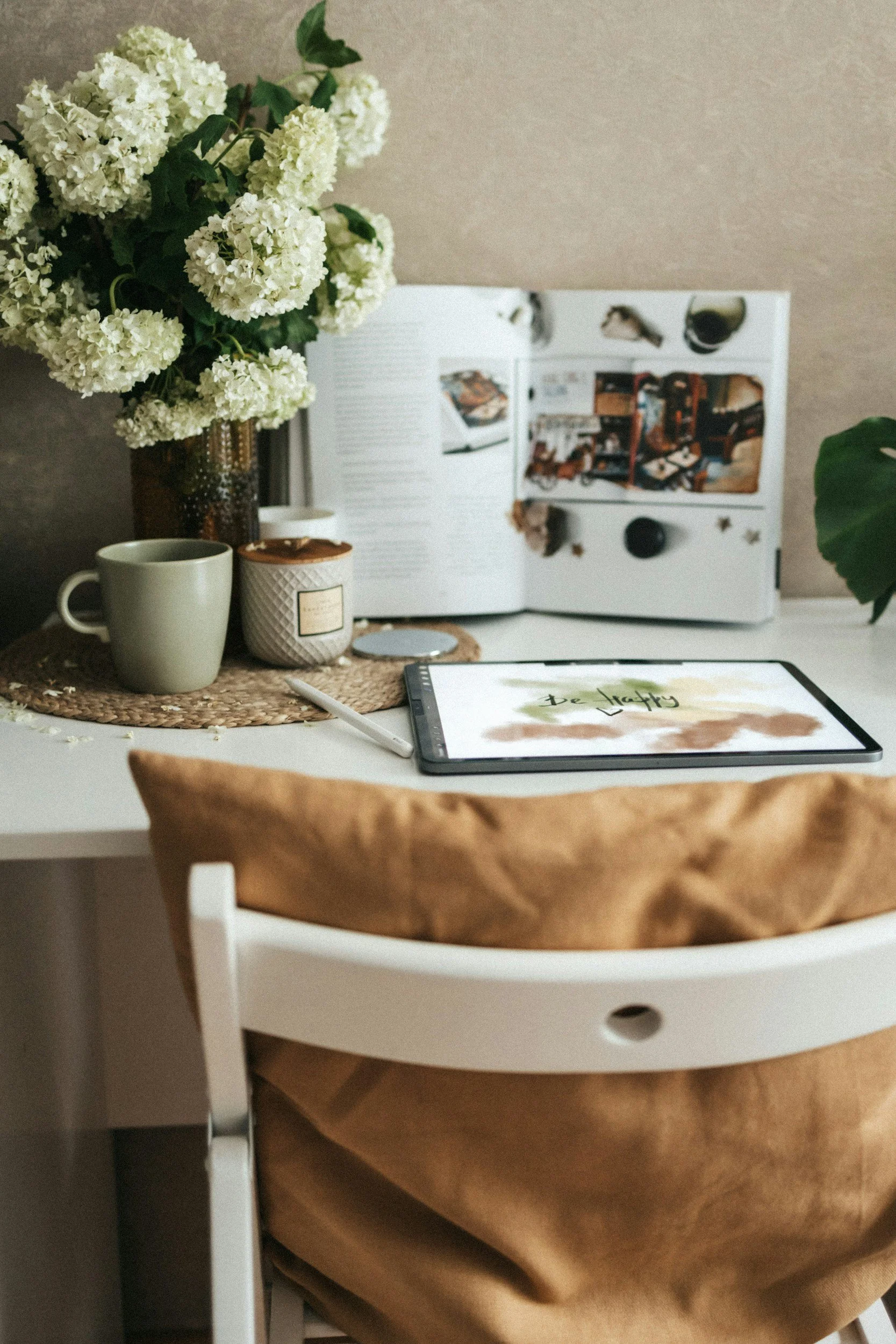A white desk with a brown paper bag, a tablet with 'Be Happy' on the screen, a green mug, a candle, a white book, and a magazine open to interior decor photos. There is a bouquet of white flowers in a brown vase and a large green leaf in the corner.