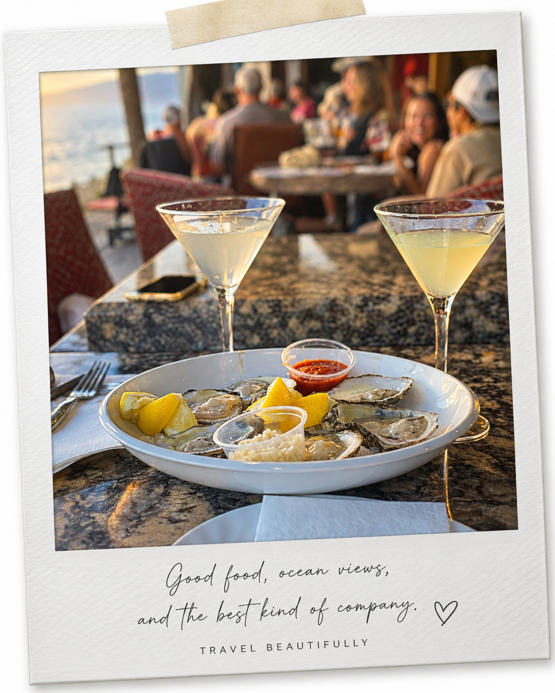 A seafood meal with oysters, lemon wedges, and condiments on a plate, two cocktails in martini glasses, and a sunset ocean view in the background where people are dining outdoors at a seaside restaurant.
