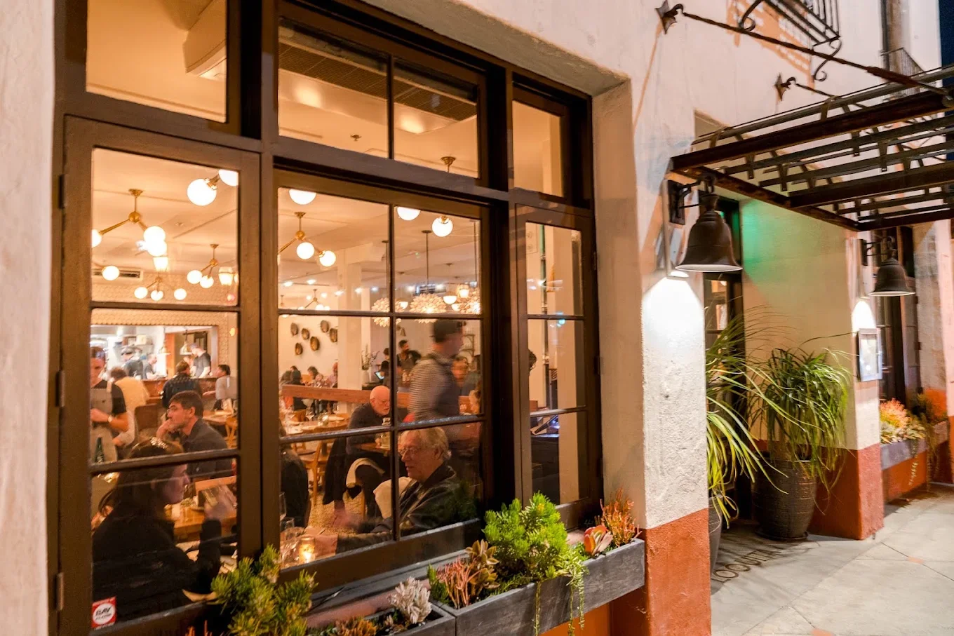 View of a busy restaurant interior through a large window, with warm lighting, diners eating and talking, and decor including plants, hanging lights, and wall ornaments.