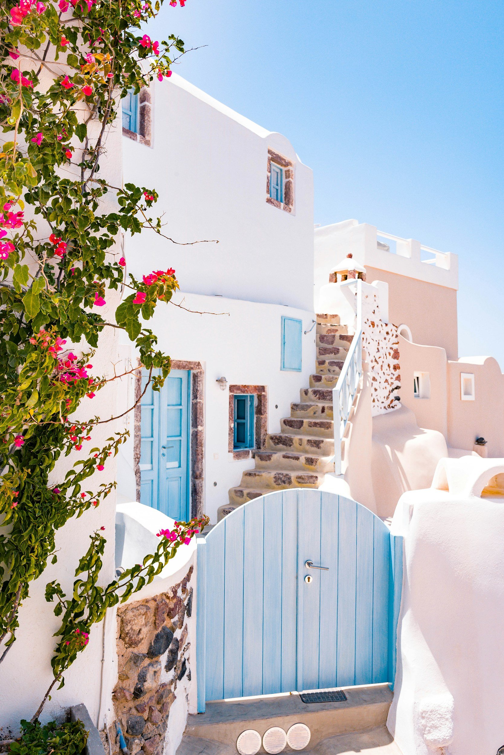 White building with blue doors and windows, stone accents, a staircase with a white railing, pink flowers climbing a wall, and a blue gate at the bottom of the stairs, under a clear blue sky.