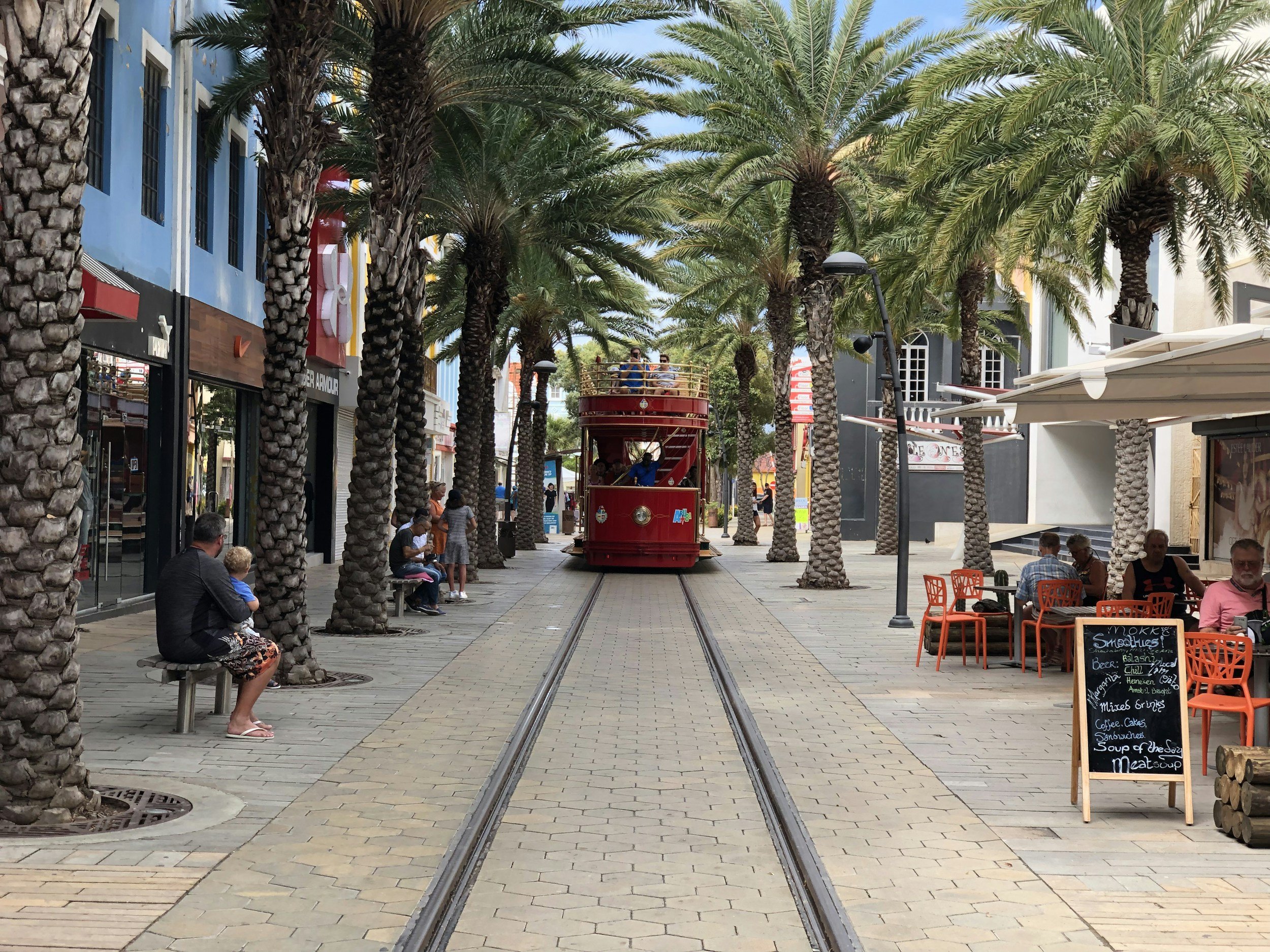 Street scene with palm trees, shops, pedestrians, and a red trolley on a track, resembling a shopping or entertainment district.
