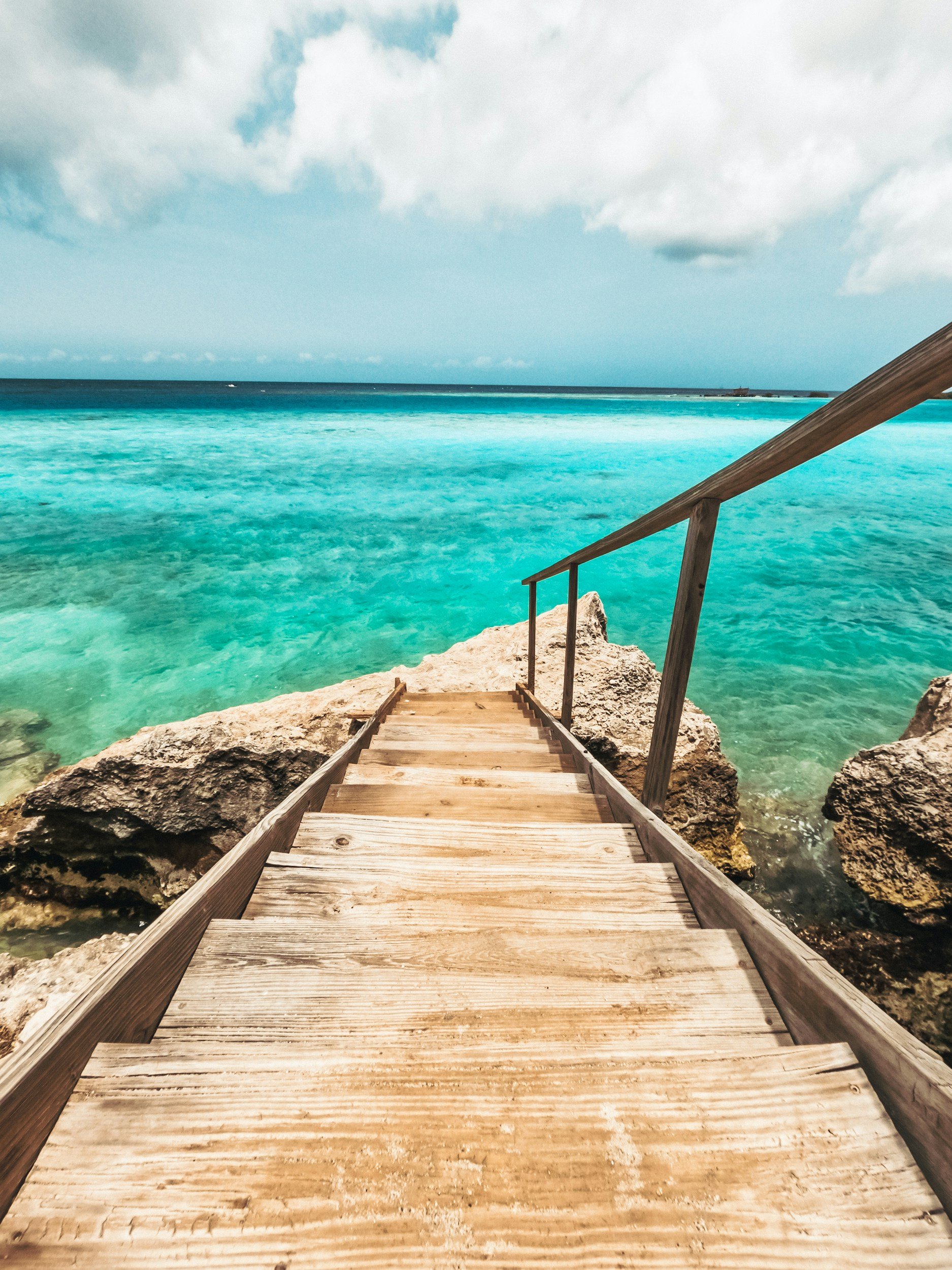Wooden steps leading down to a rocky shoreline with turquoise water, overlooking the ocean, under a partly cloudy sky.