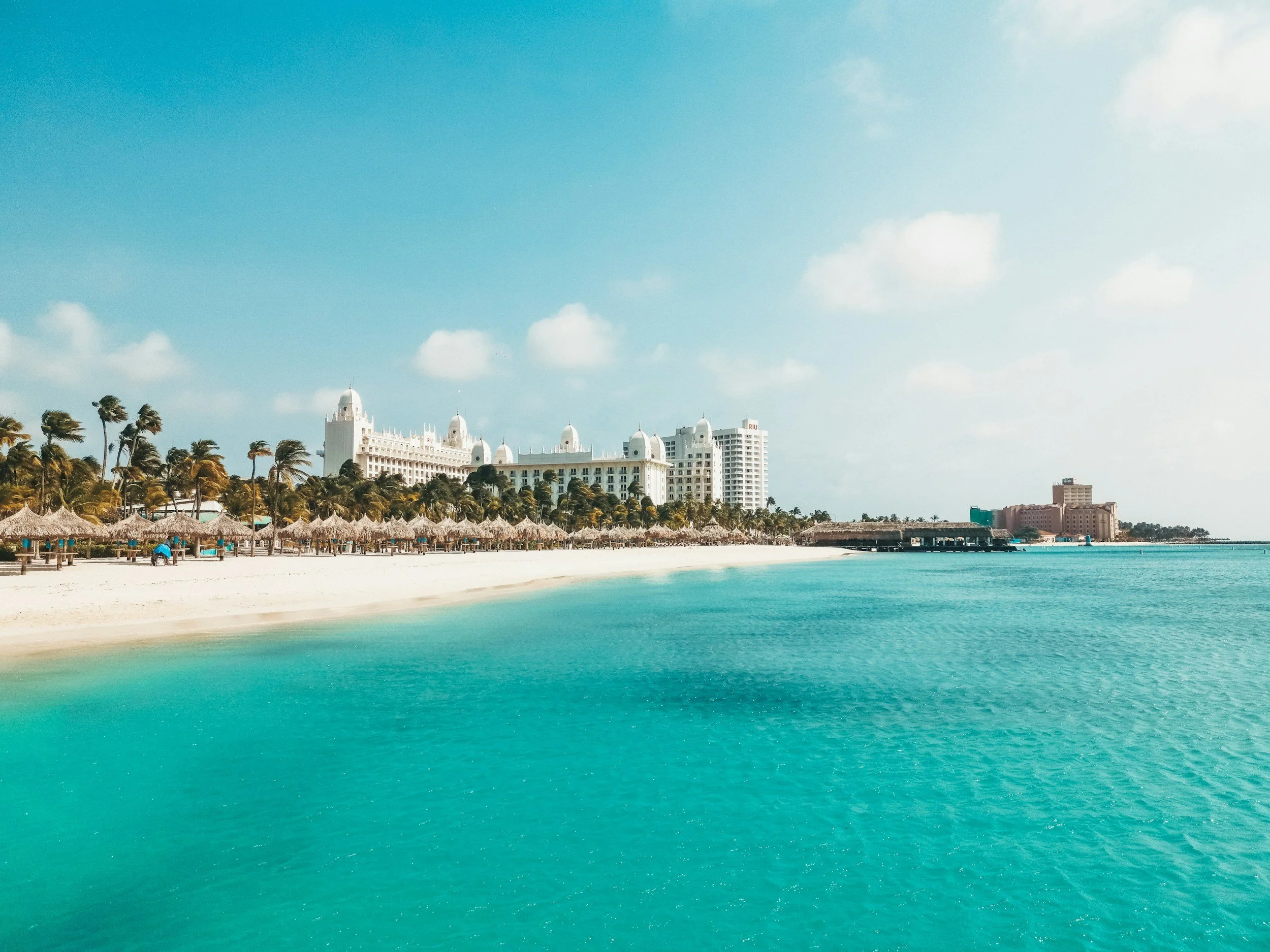 Photo of a beach with straw umbrellas and lounge chairs, palm trees, and a large white hotel building in the background under a partly cloudy sky.