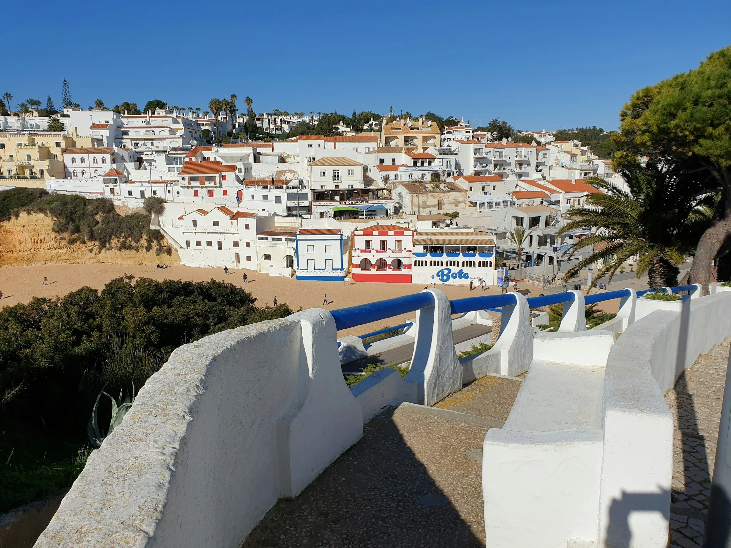 View of a hillside town with white buildings and red-tiled roofs under a clear blue sky, with palm trees visible on the hilltop and a beach with a few people in the foreground.