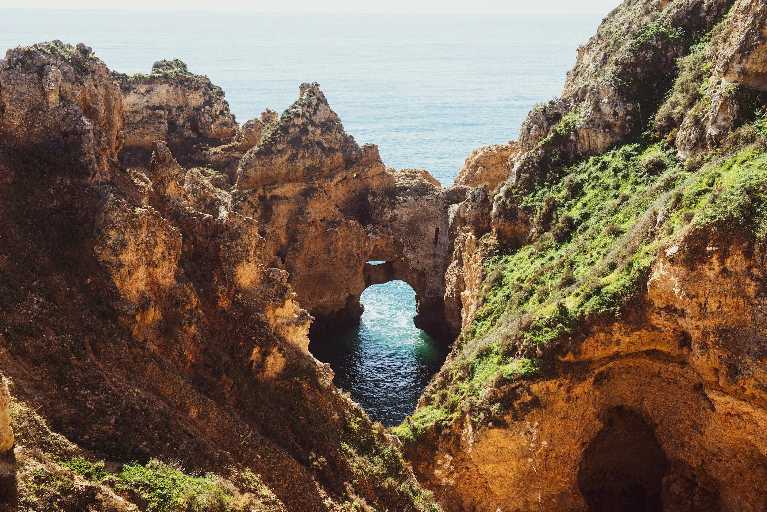 Cliffside with natural rock arch letting water through, overlooking the ocean with blue sky and light clouds.