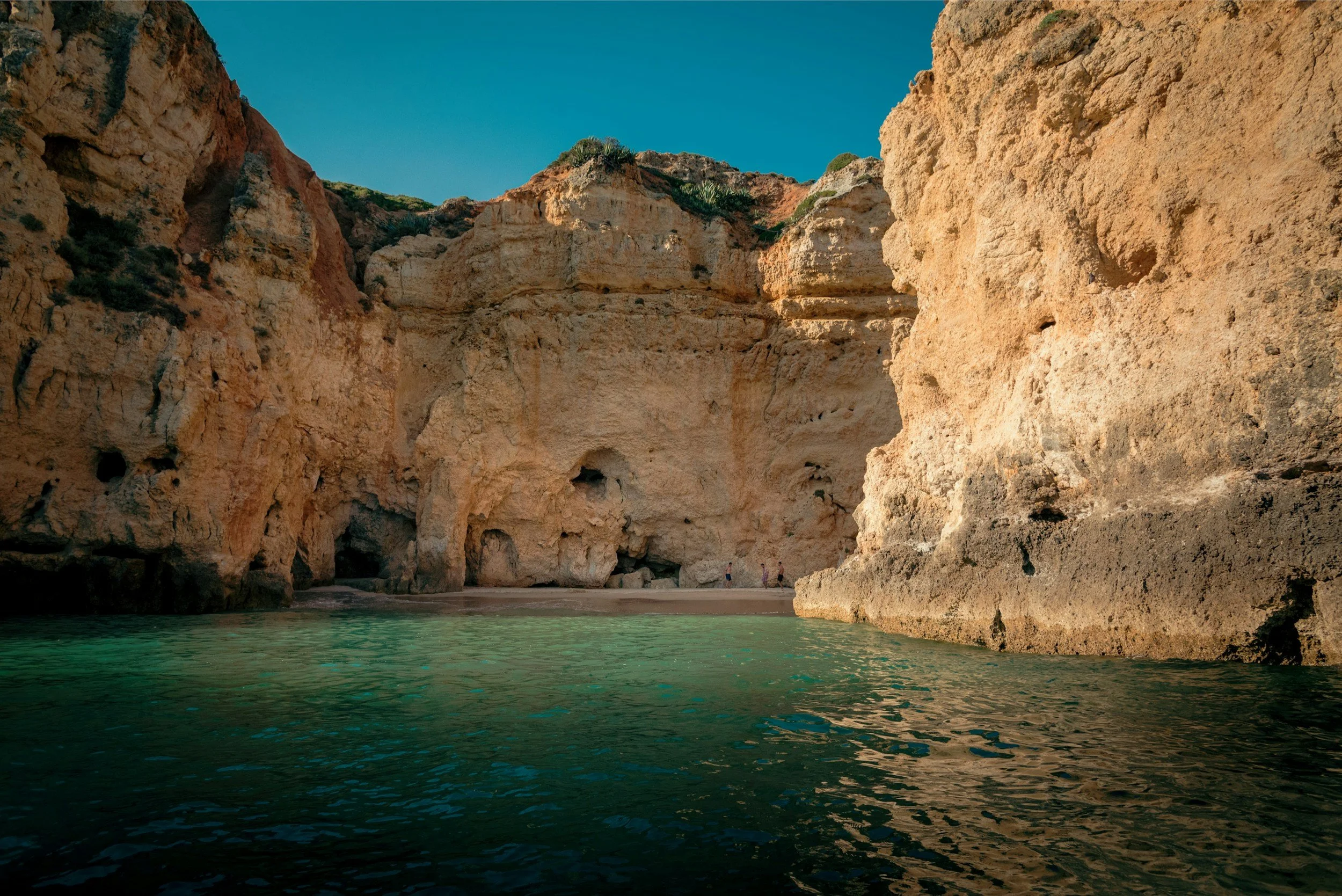 A scenic coastal landscape with large beige and reddish-orange cliffs rising from the turquoise water, and a small beach at the base with a few people standing near the shoreline under a clear blue sky.